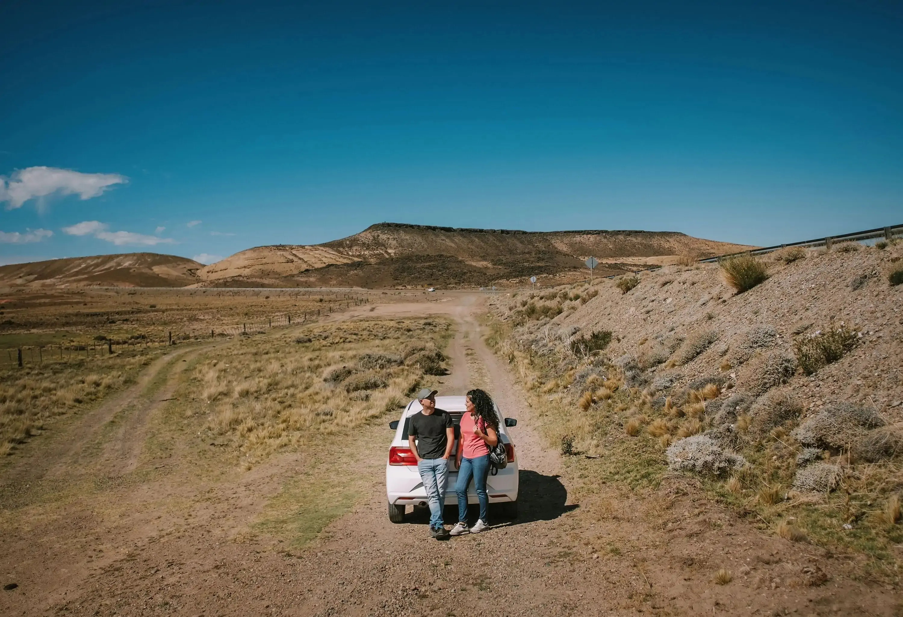 A man and woman couple are standing in front of a car. In the background you can see the Patagonian desert and its routes.
