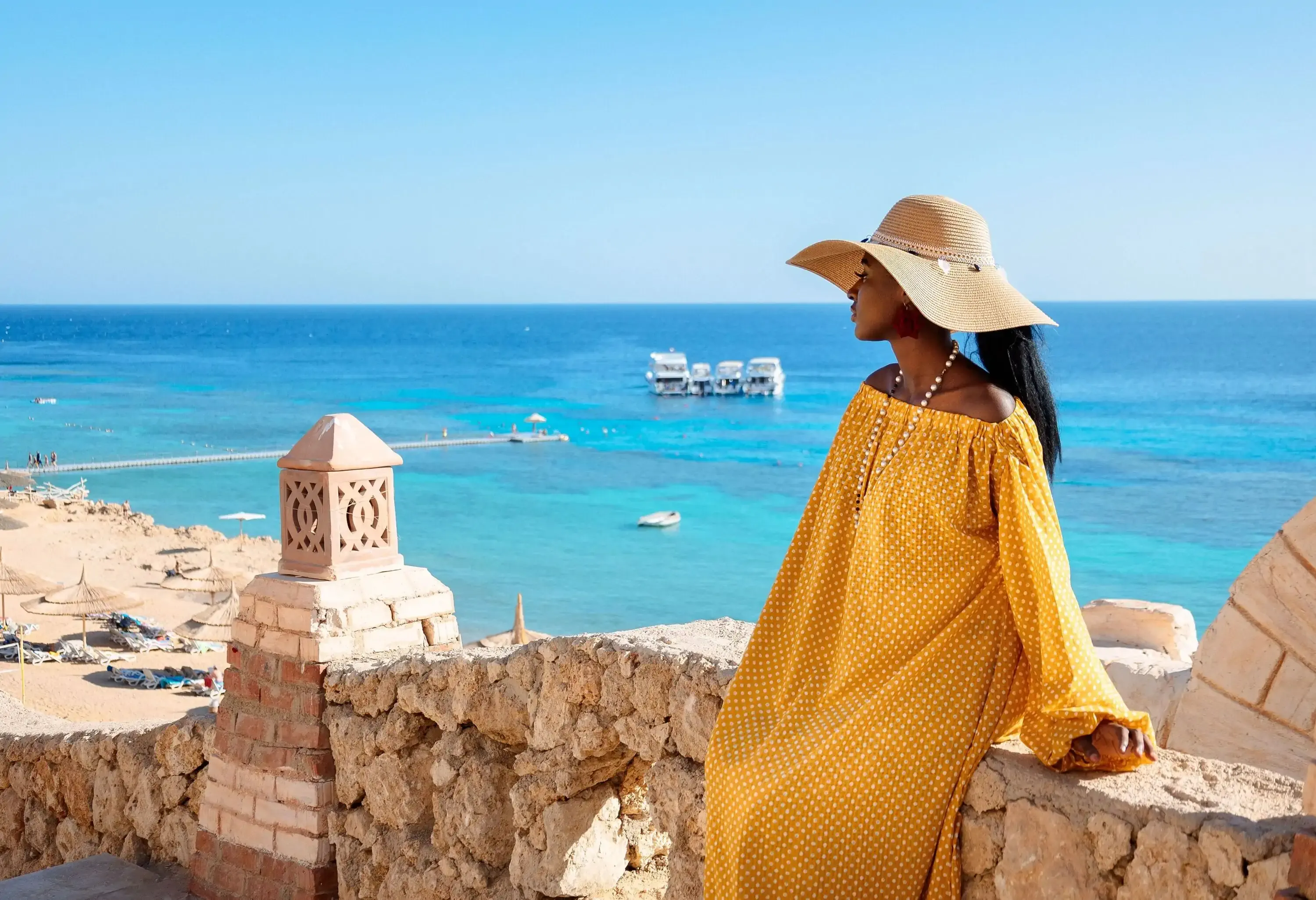 Woman in yellow dress with floppy hat standing by turquoise ocean