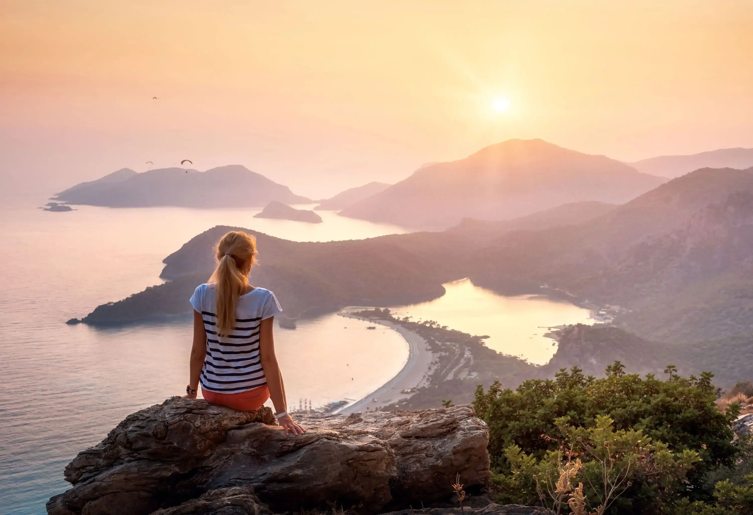 A woman sitting on a rock atop a hill, watching the sun set over the mountains across the sea.