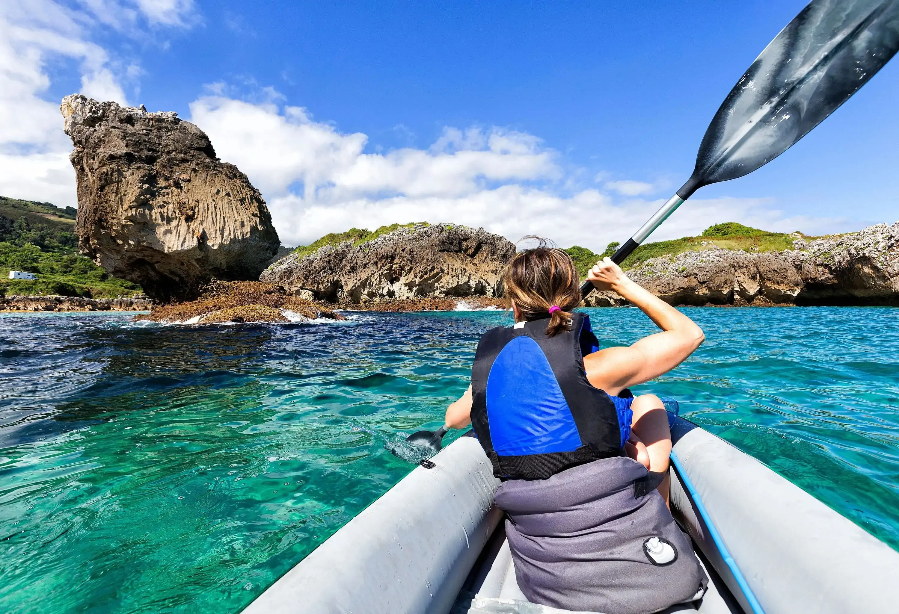 A woman paddles a kayak in the ocean, approaching a lush rocky shore.