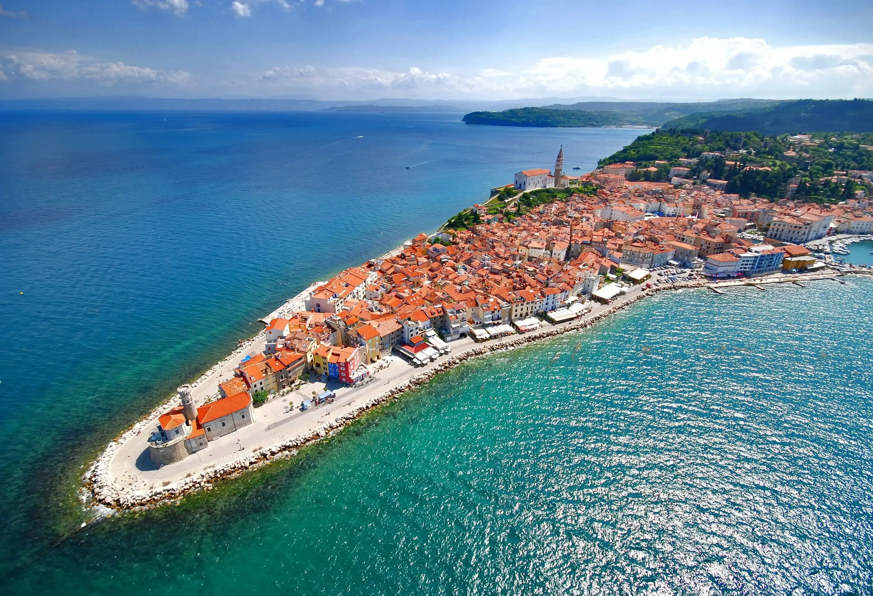 An aerial view of a picturesque coastal town on a narrow peninsula, featuring tightly packed buildings with red-tiled roofs, a historic lighthouse, and a church tower, surrounded by the deep blue sea.