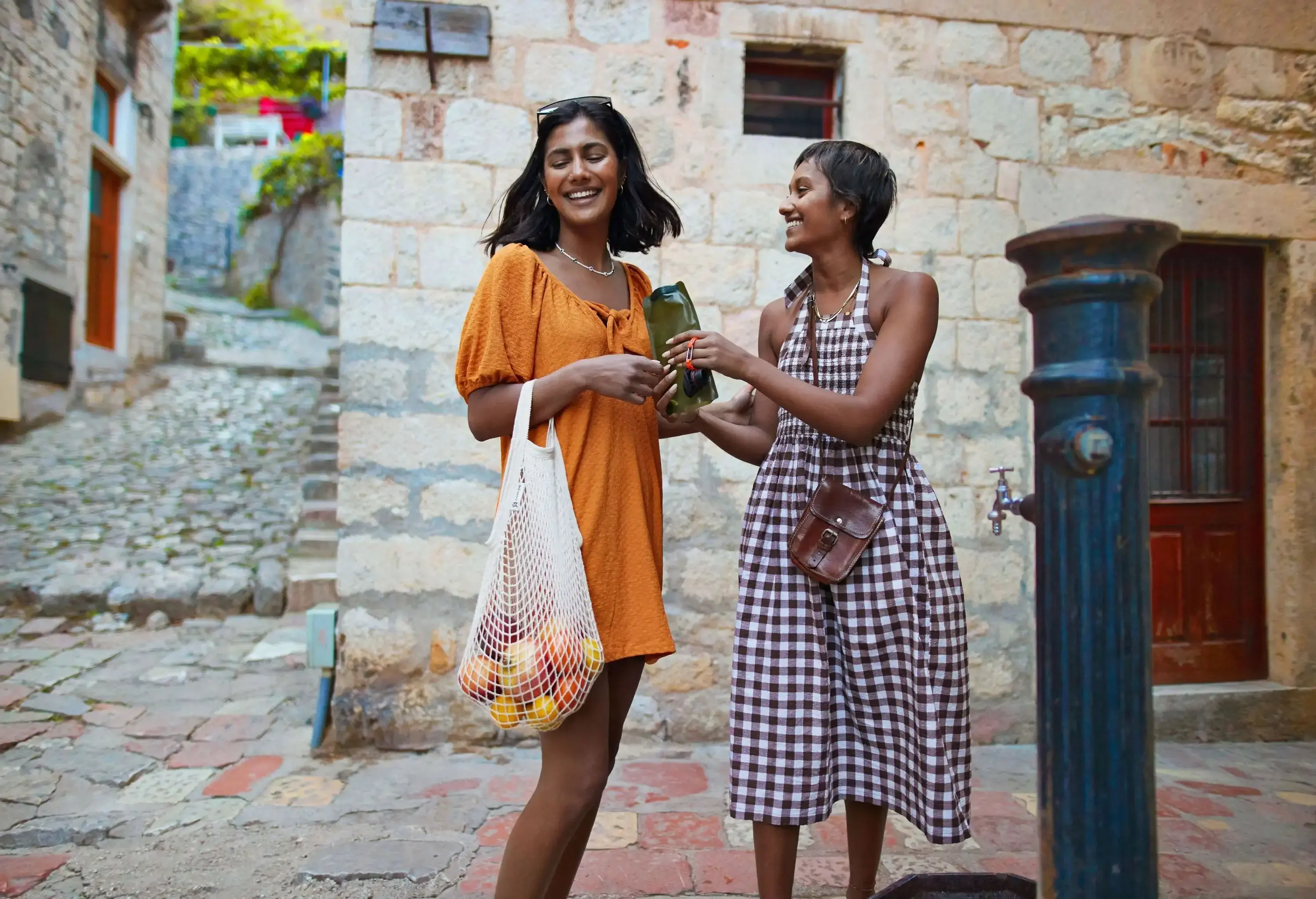 Two young women laughing and sharing a drink in a charming old stone-paved alley, with one carrying a mesh bag filled with fresh fruit, surrounded by rustic architecture.