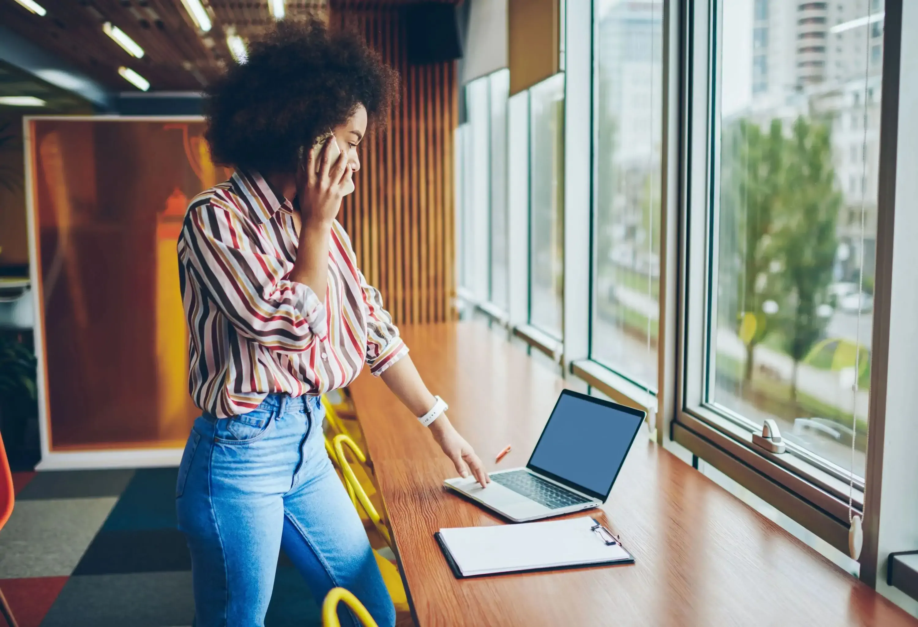 A woman with beautiful afro hair, standing and talking on her phone while looking at her laptop on the table.