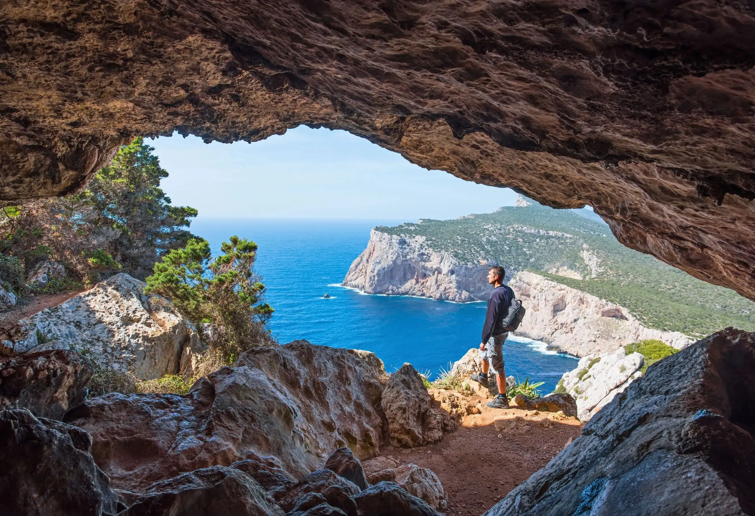 A male hiker standing along the edge outside of a cave overlooking the blue sea.