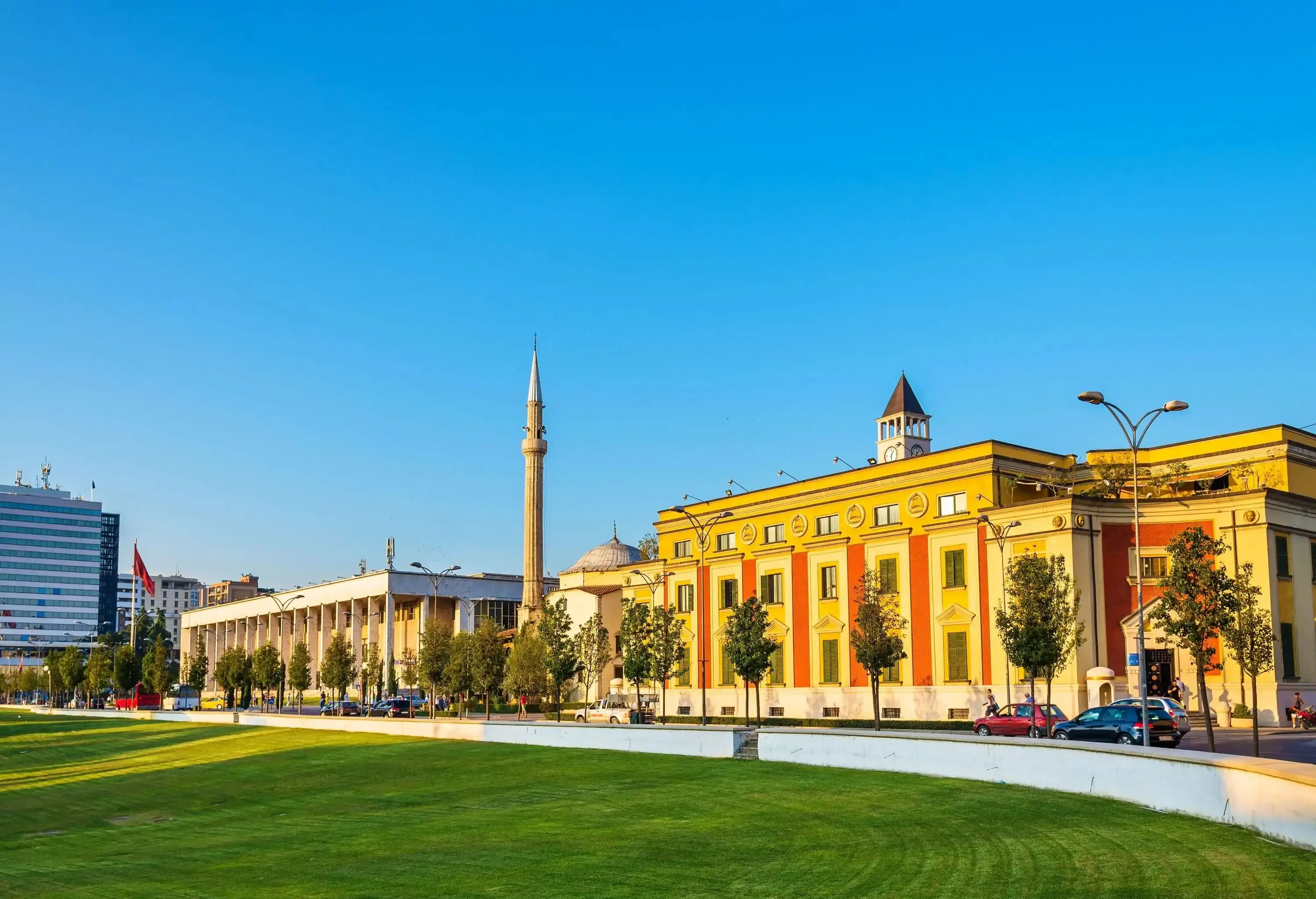 A wide tree-lined green lawn with views of a pointed monument in the middle of city buildings illuminated by the sun.