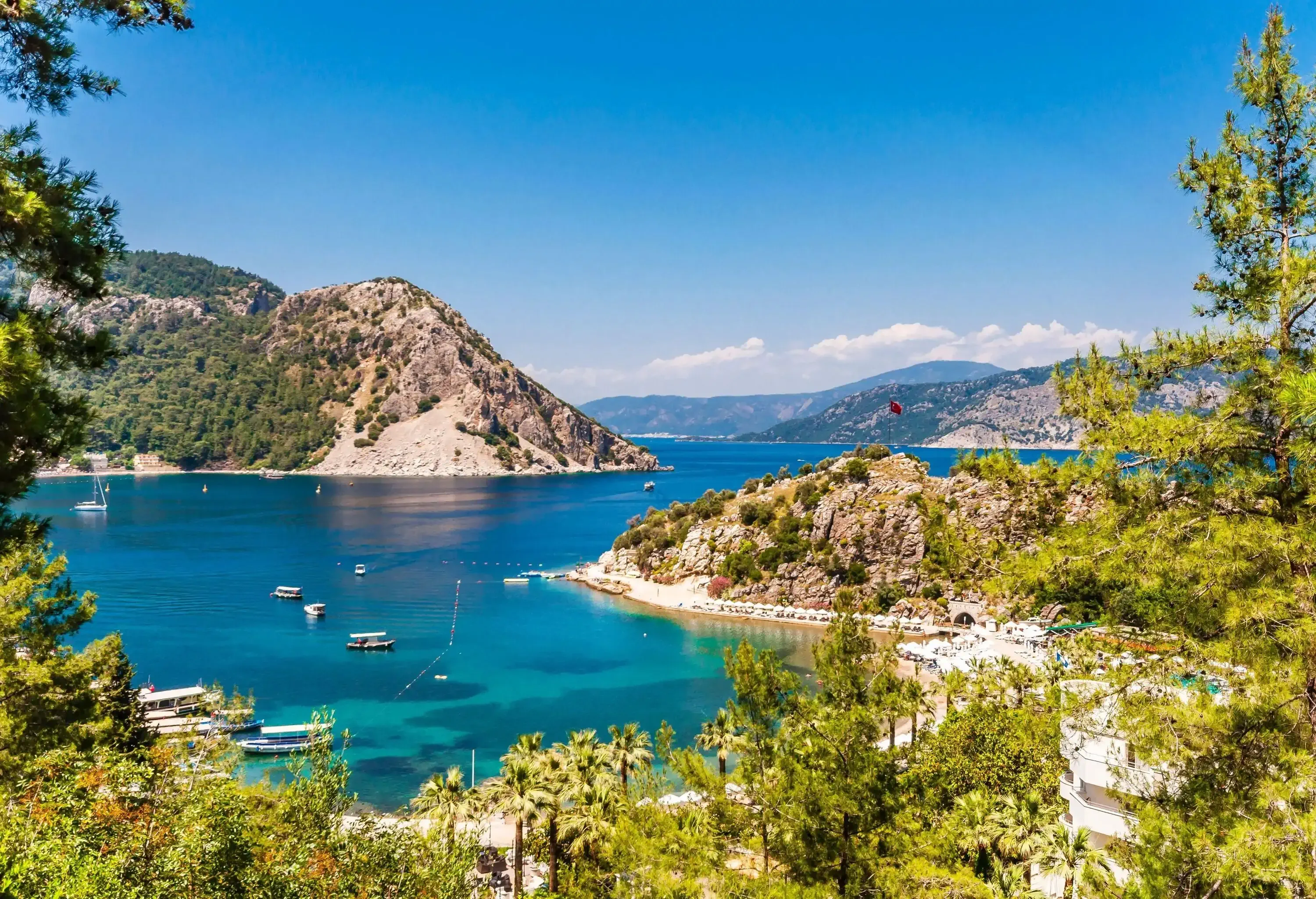 A picturesque coastal landscape featuring a turquoise bay surrounded by lush green hills, rocky cliffs, and a sandy beach with sun loungers, framed by pine trees under a clear blue sky.