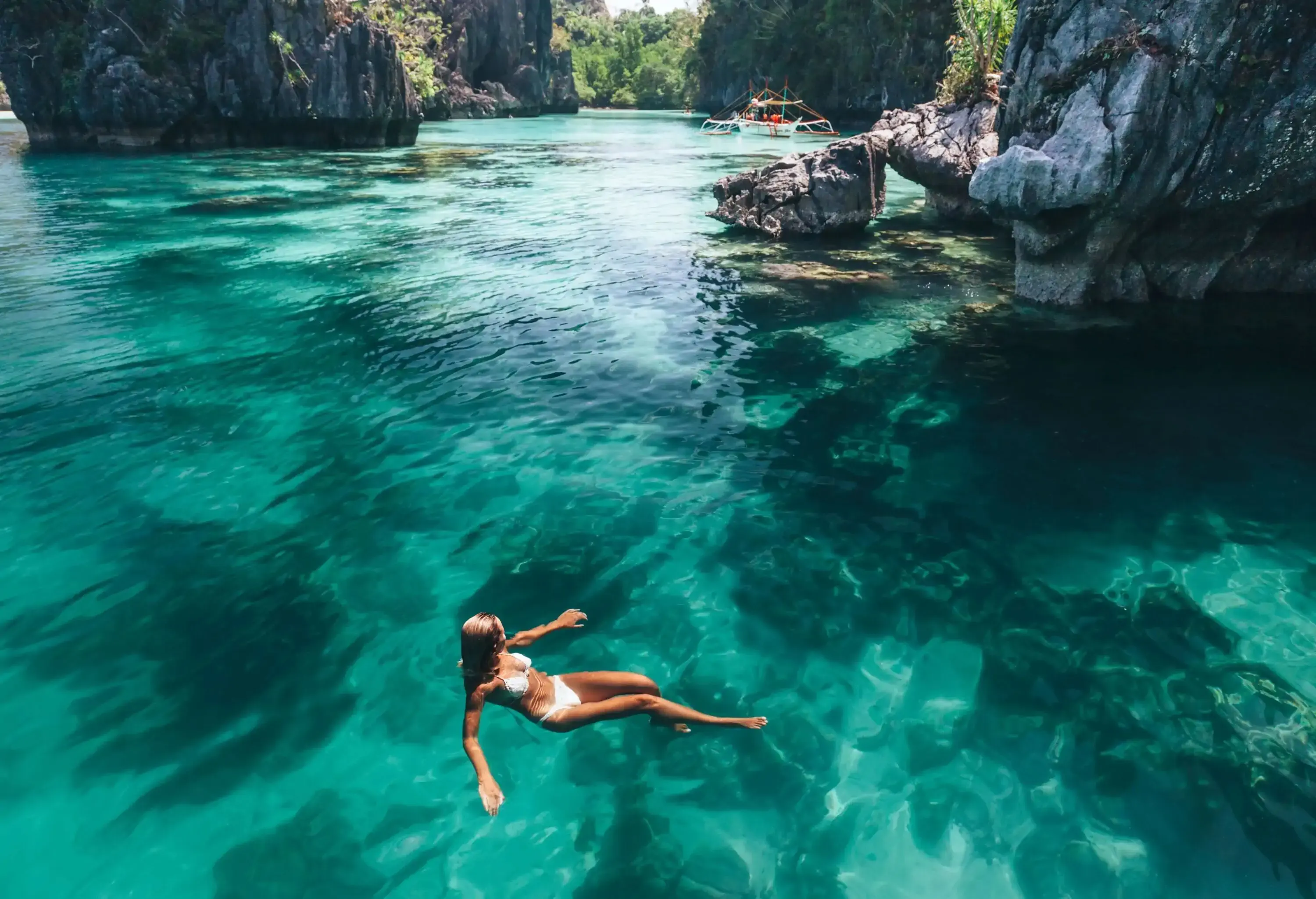 Aerial view of a woman in a white bikini floating on a clear water sea overlooking the towering blackish rock formations.