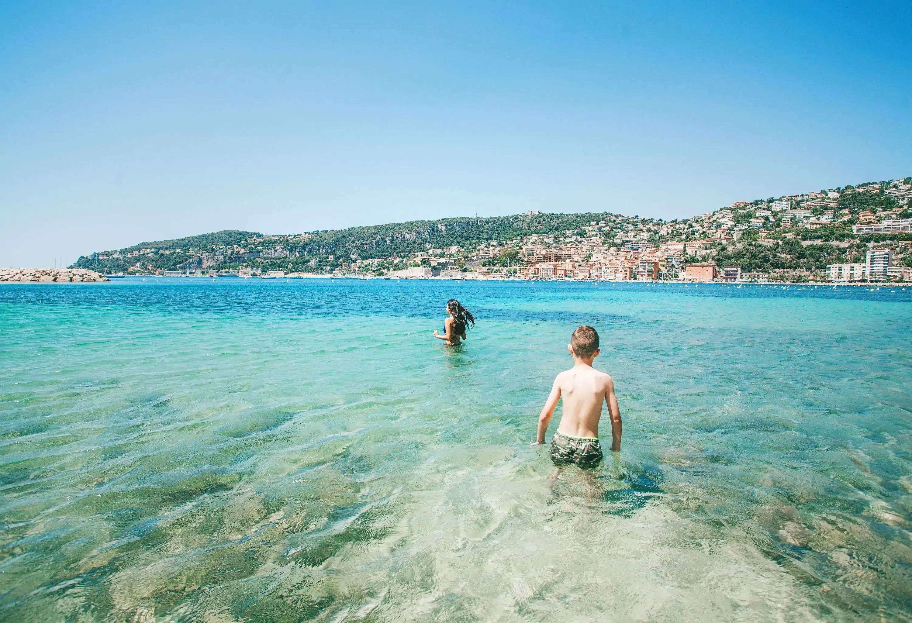 Two young individuals on a clear water beach overlooking the coastal hillside cityscape.