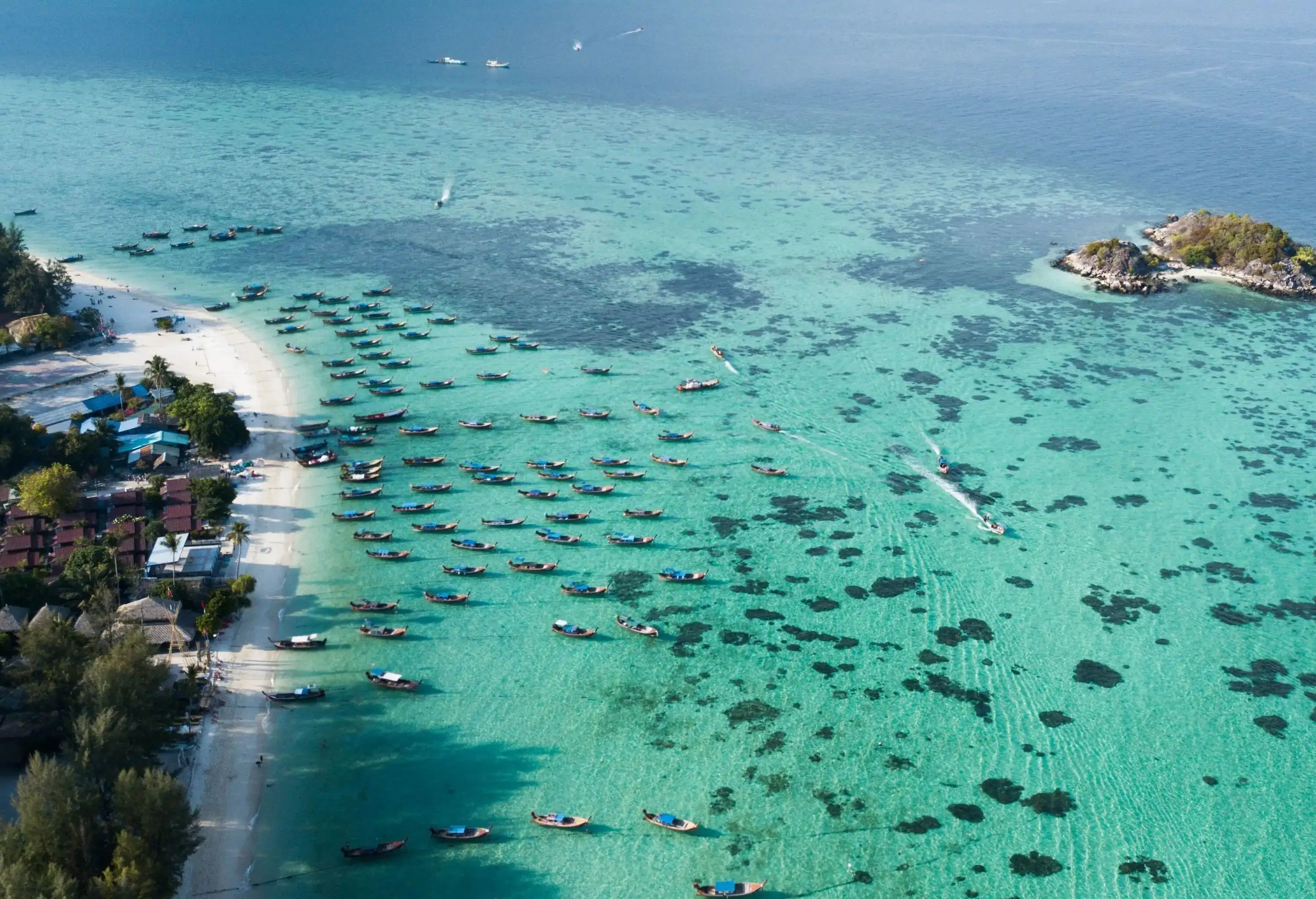 Aerial view of a clear water sea dotted with fishing boats and the white sand shore surrounded by tall trees and houses.