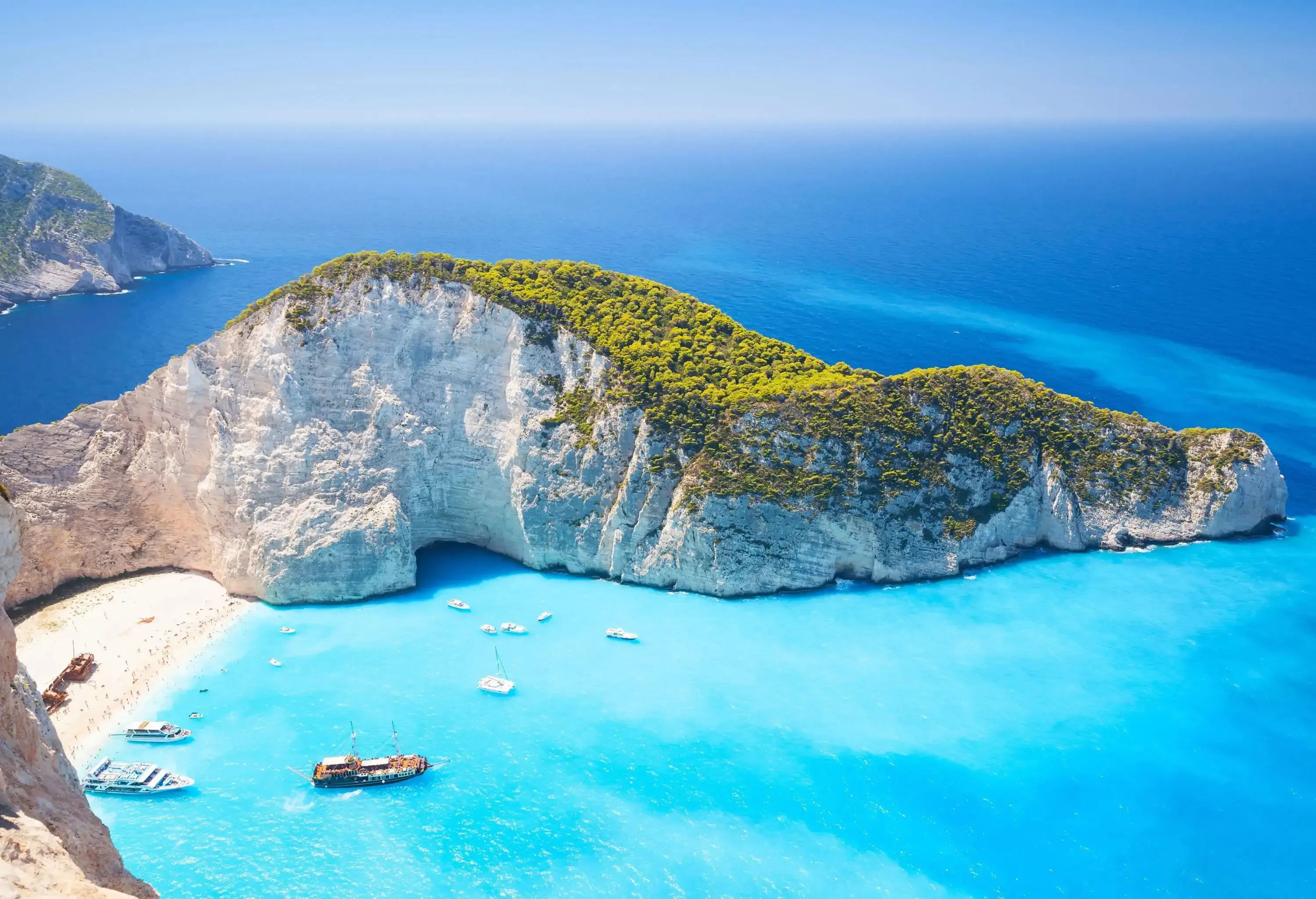 A white steep cliff with lush foliage beside a cove with docked boats.