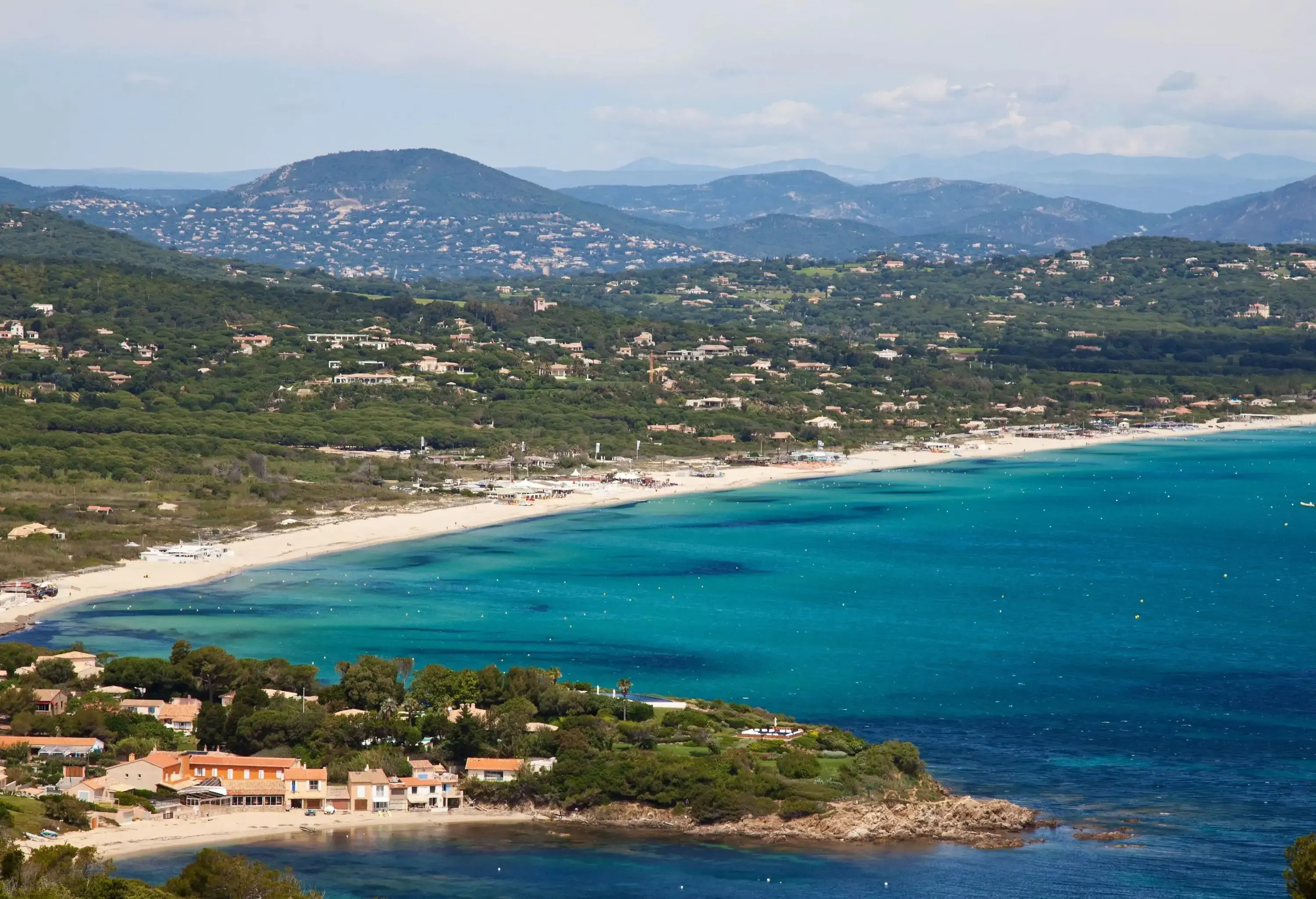 A white beach with neighbourhoods perched on forested hills and sandy coastlines. 