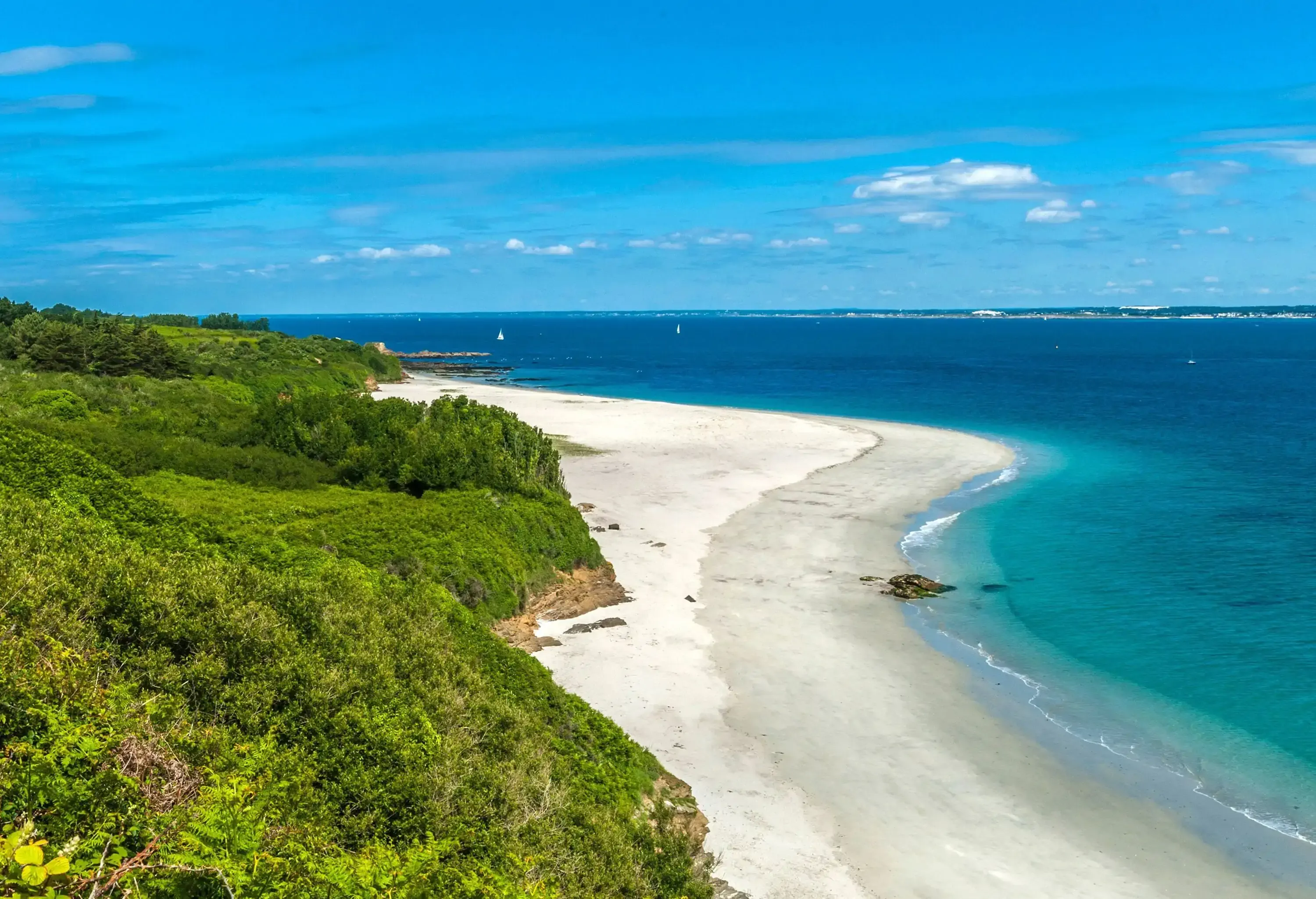 A lush seaside hill and the white sand shore of the blue water beach against the clear blue sky.