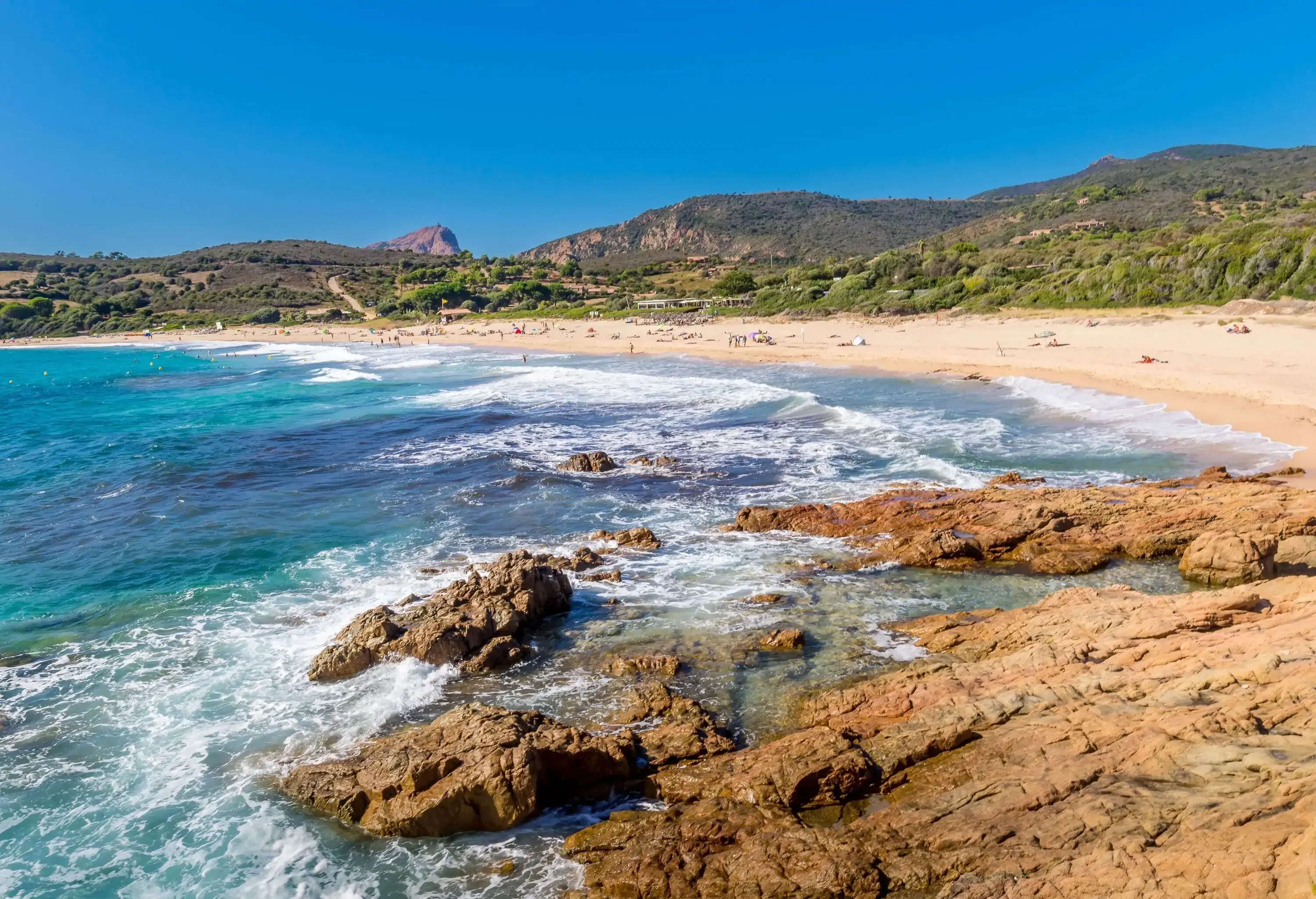A blue water wavy beach, a sandy shore, a lush hillside and a lush mountain range.