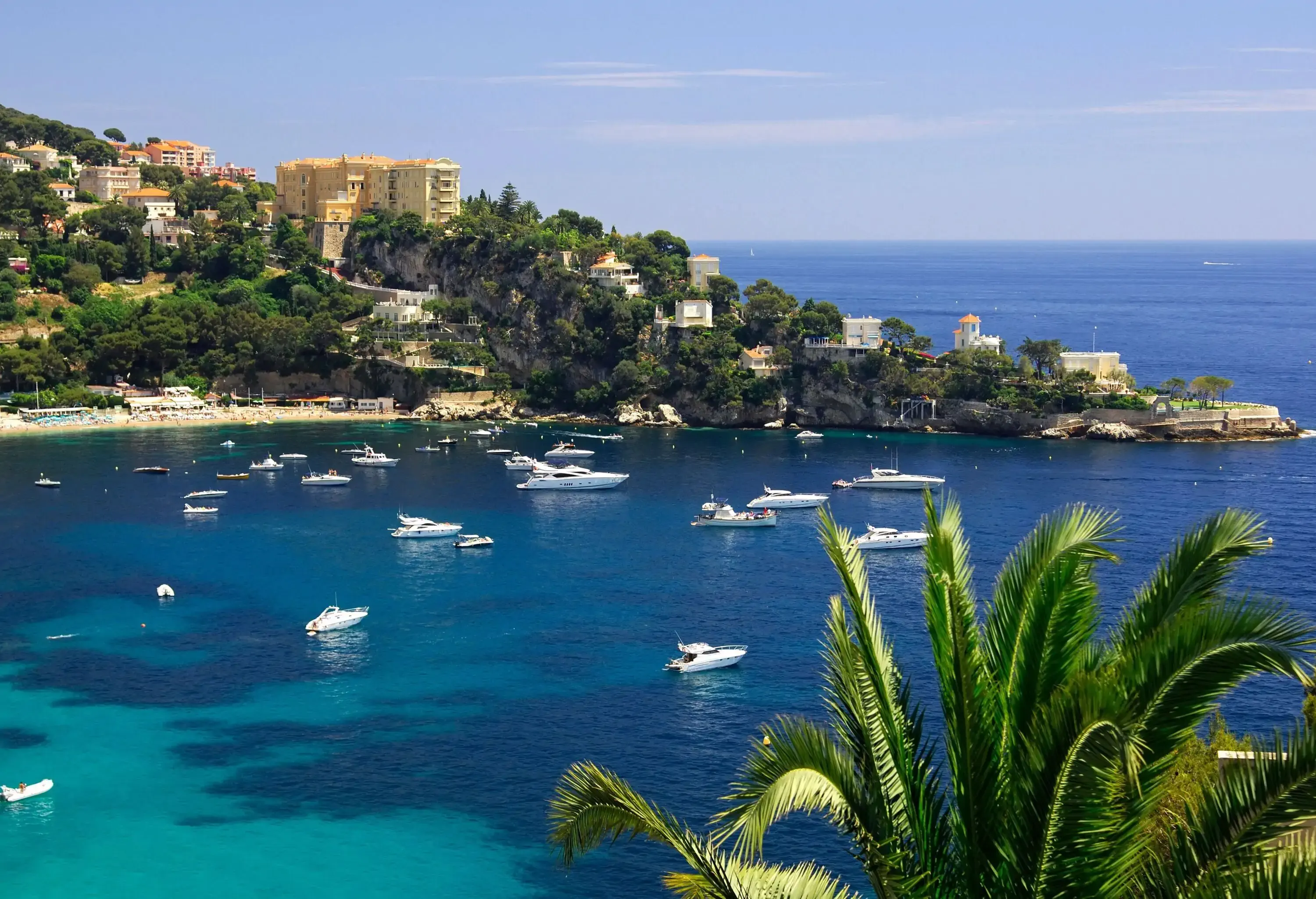 Boats moored on an azure bay alongside a forested hill dotted with houses and buildings.