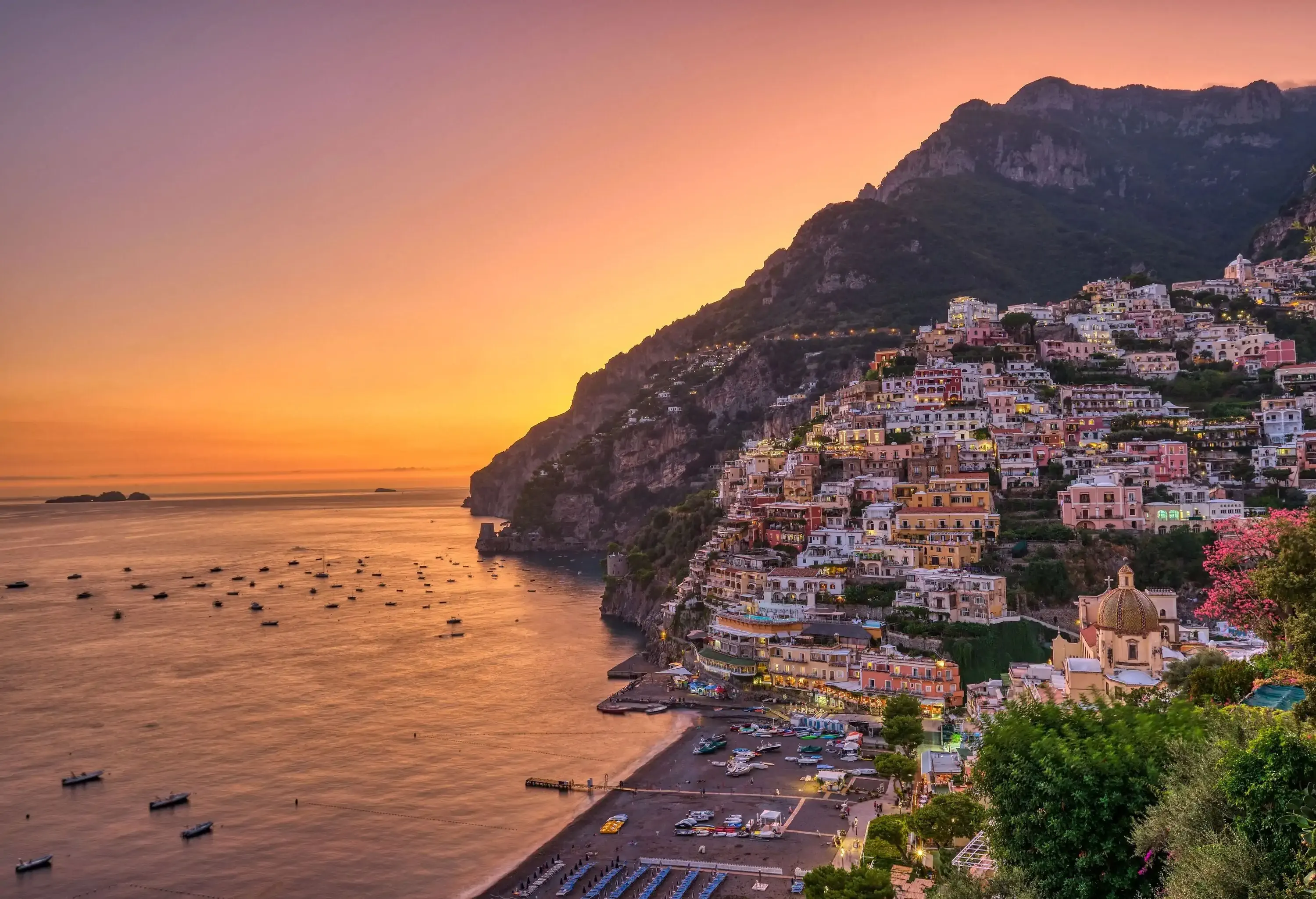 A colourful coastal village perched on the mountainside with several small moored boats at sunset.
