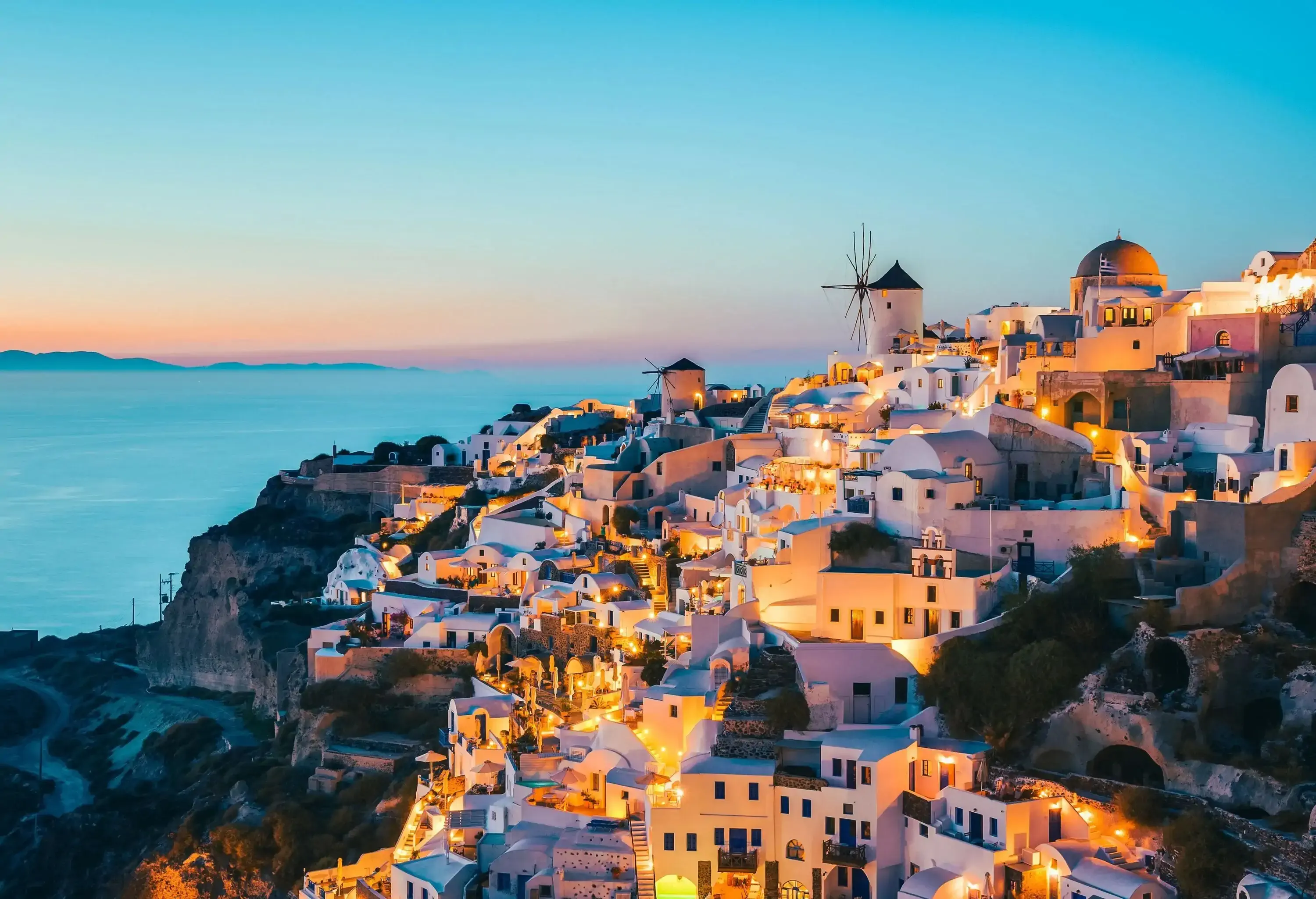 A brightly lit coastal town with traditional white-washed houses situated on a craggy hill overlooking the scenic sea at dusk.