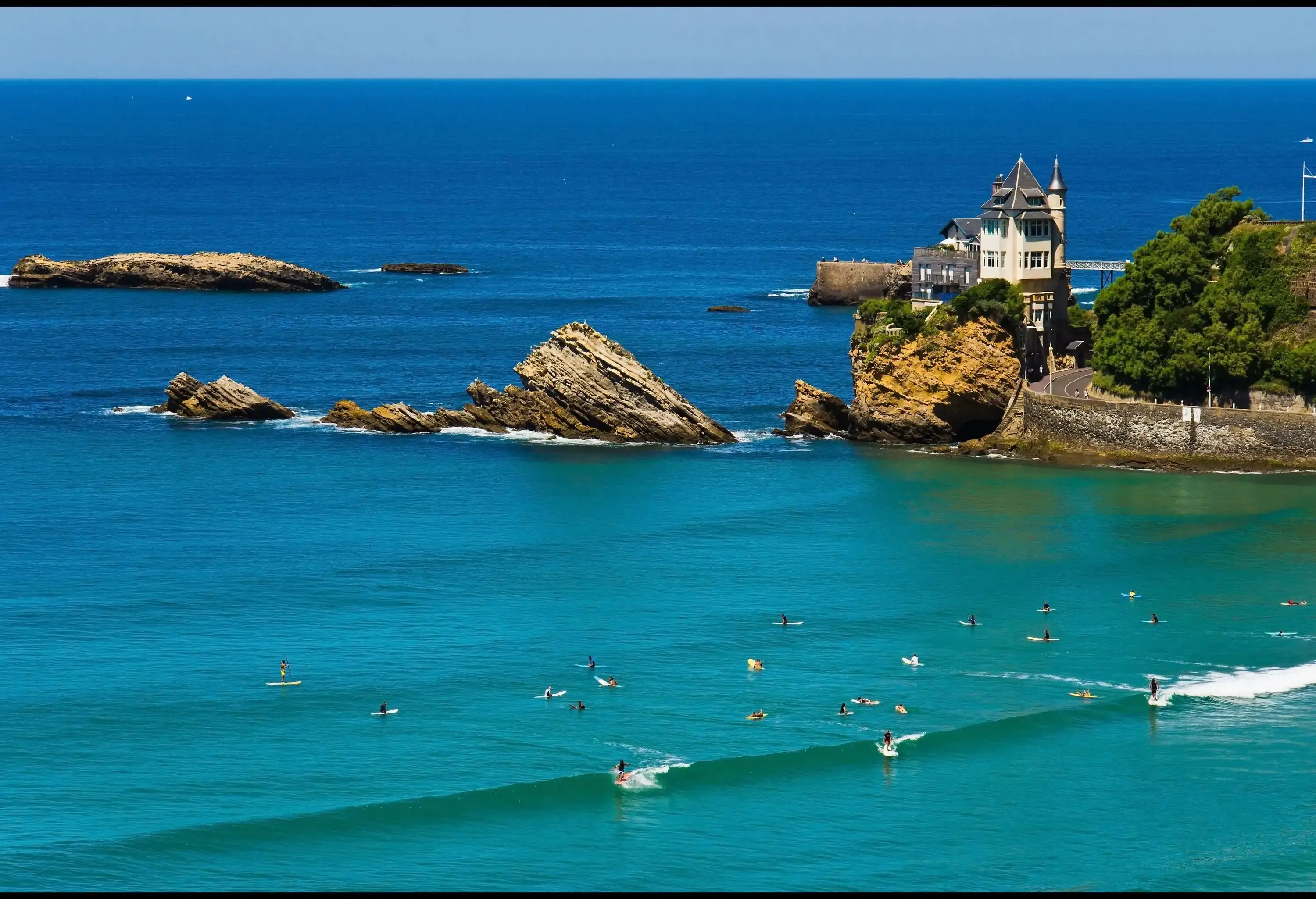 People on surfboards ride small waves near a big rock sticking out of the water next to a house built on the edge.