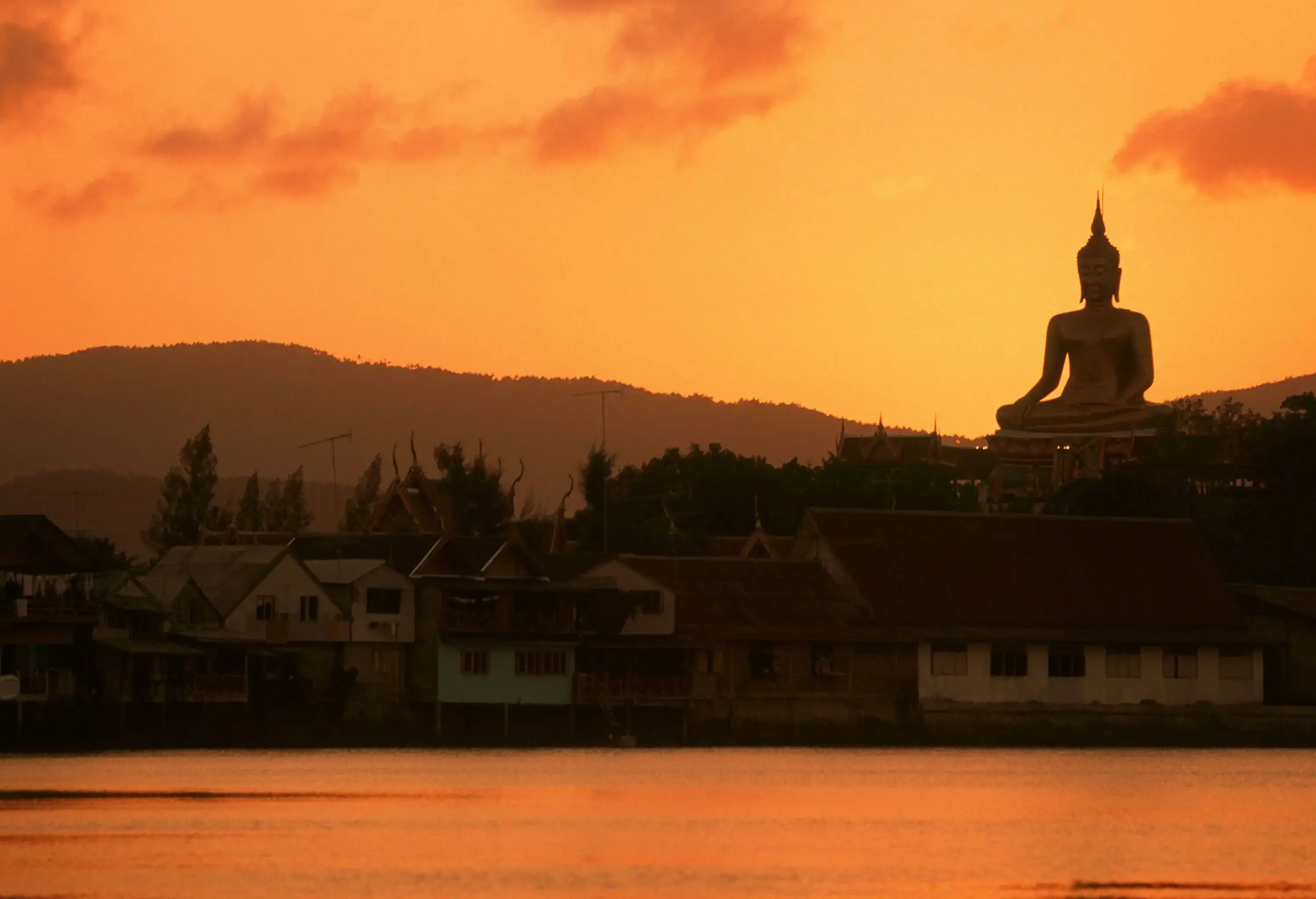 Silhouette of a big statue of a sitting Buddha overlooking a coastal village under an orange sky.
