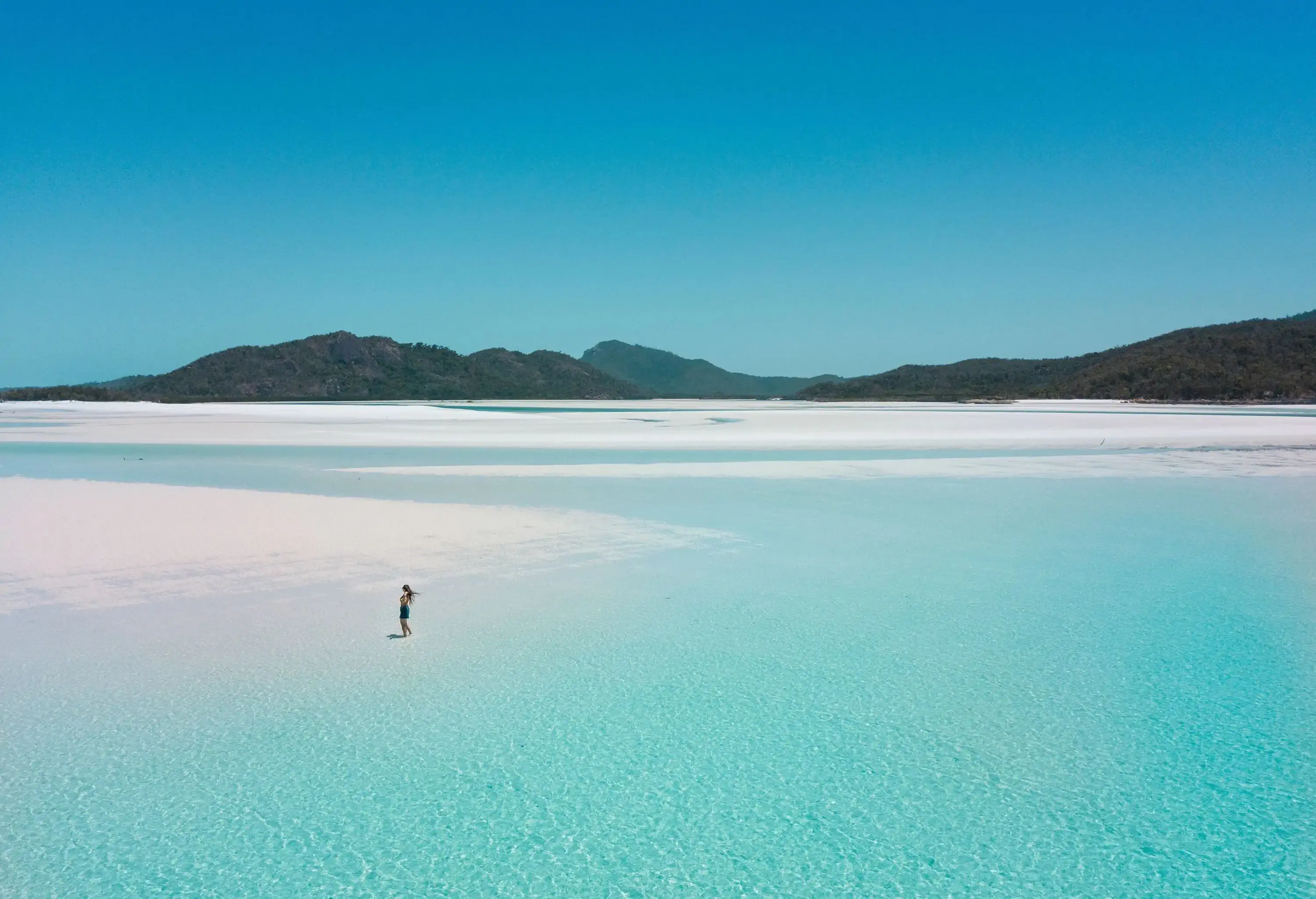 A woman on an astonishing beach with dazzling turquoise shallow water surrounded by lush mountains.