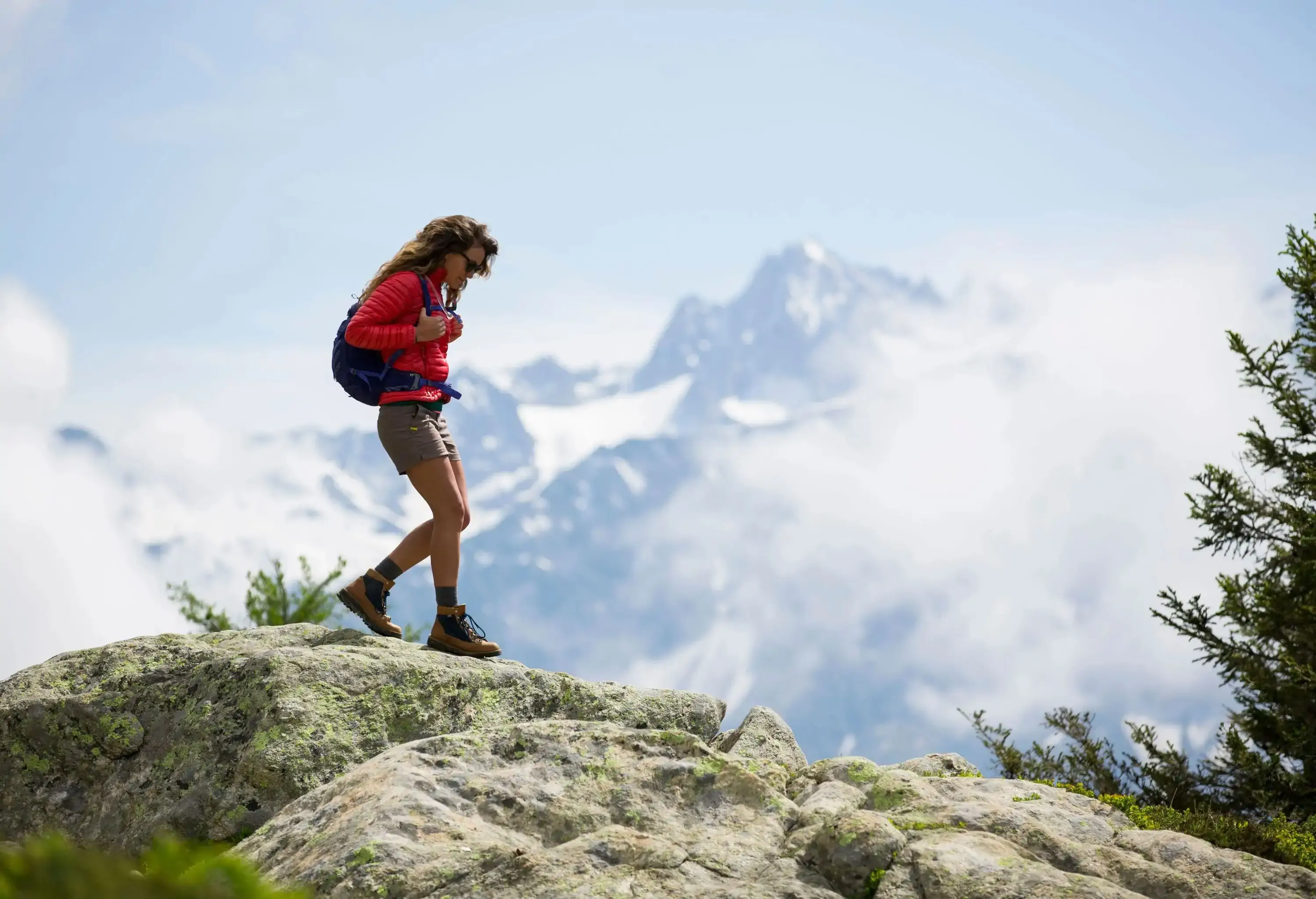 A woman with a red jacket and rucksack stands on a rock overlooking mountains covered in snow and a sea of ​​clouds.