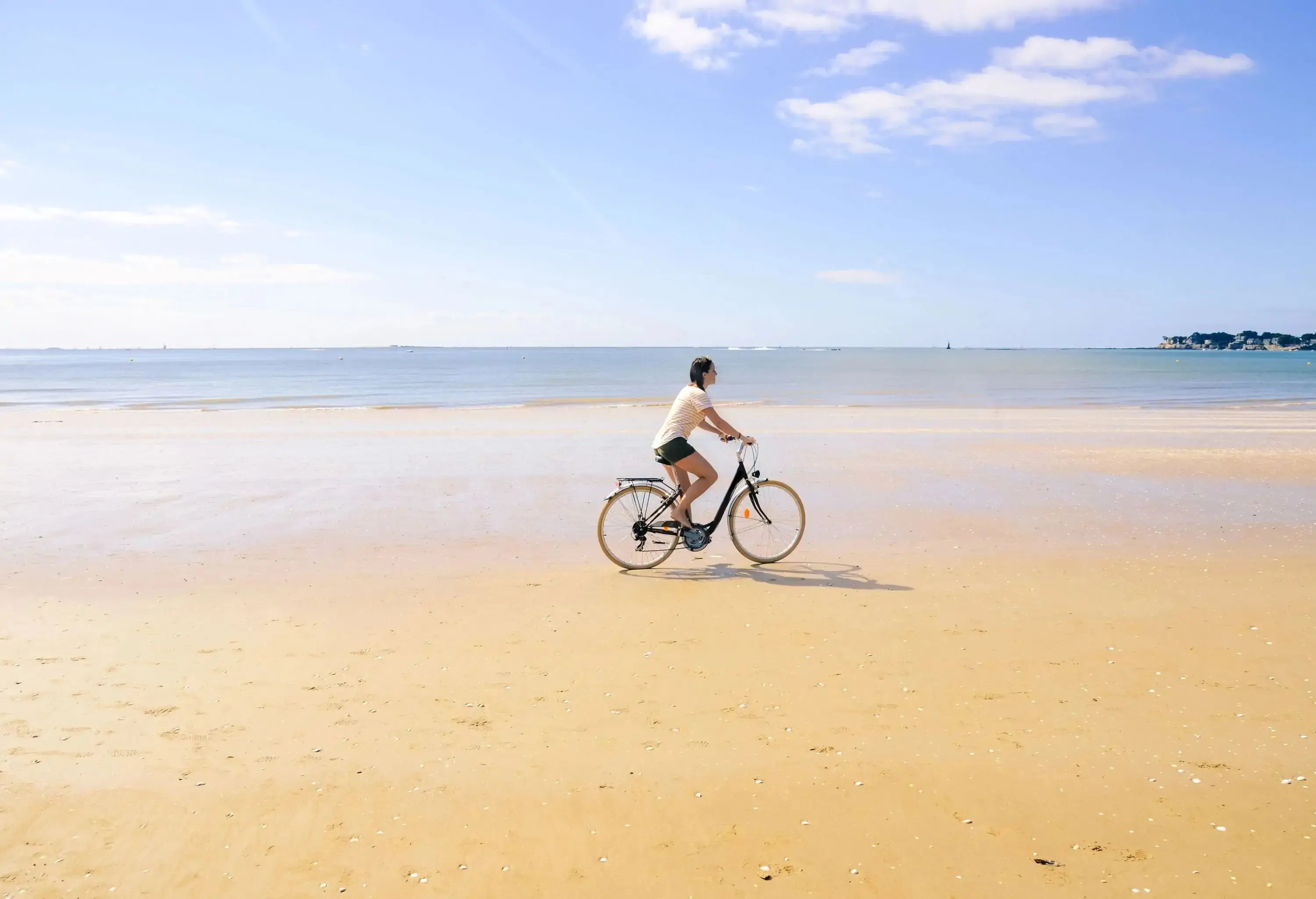 A girl is riding a bike on golden sand with a view of the calm ocean in the background.