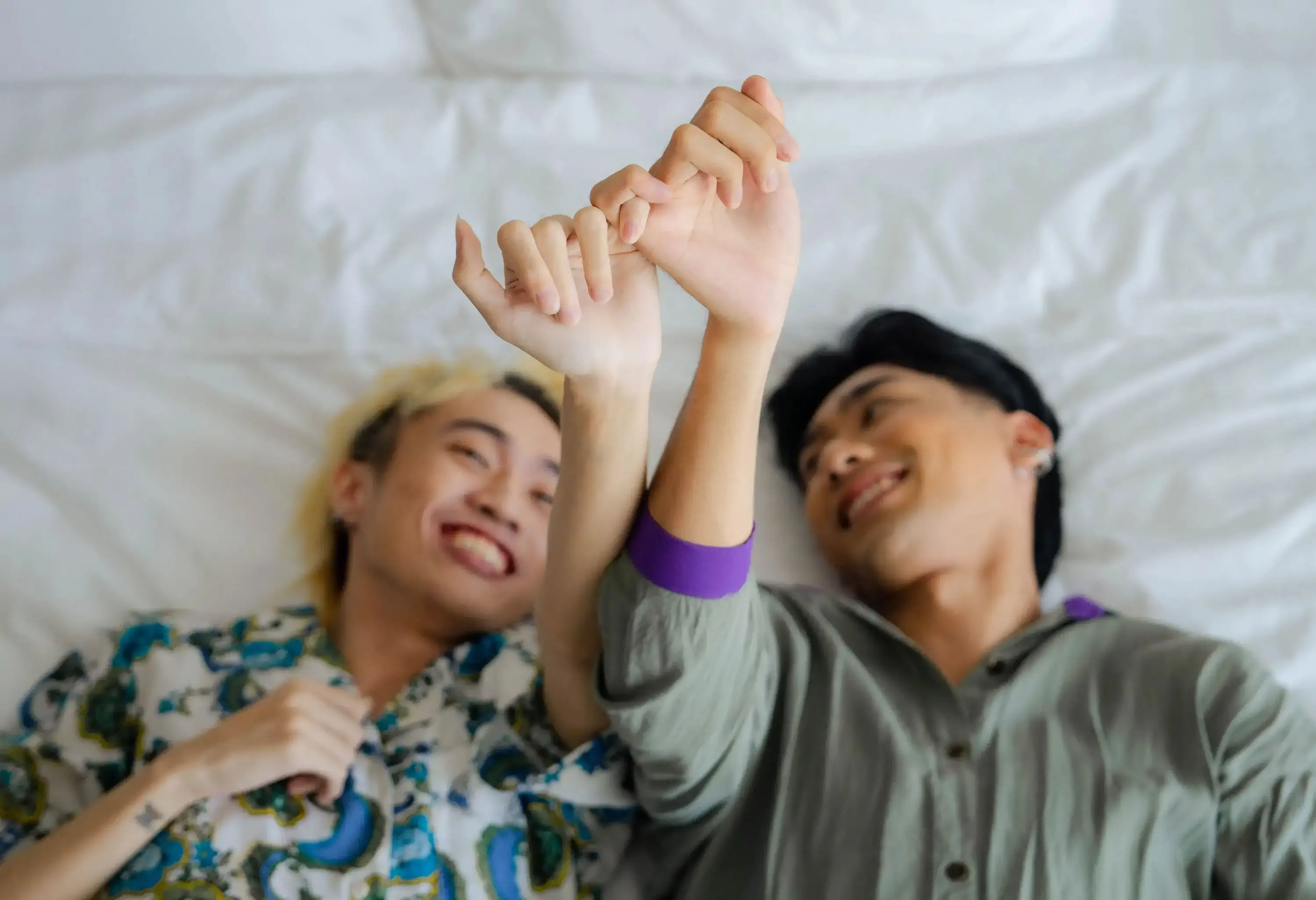 A male couple lies on the bed as they lock their pinkies together.