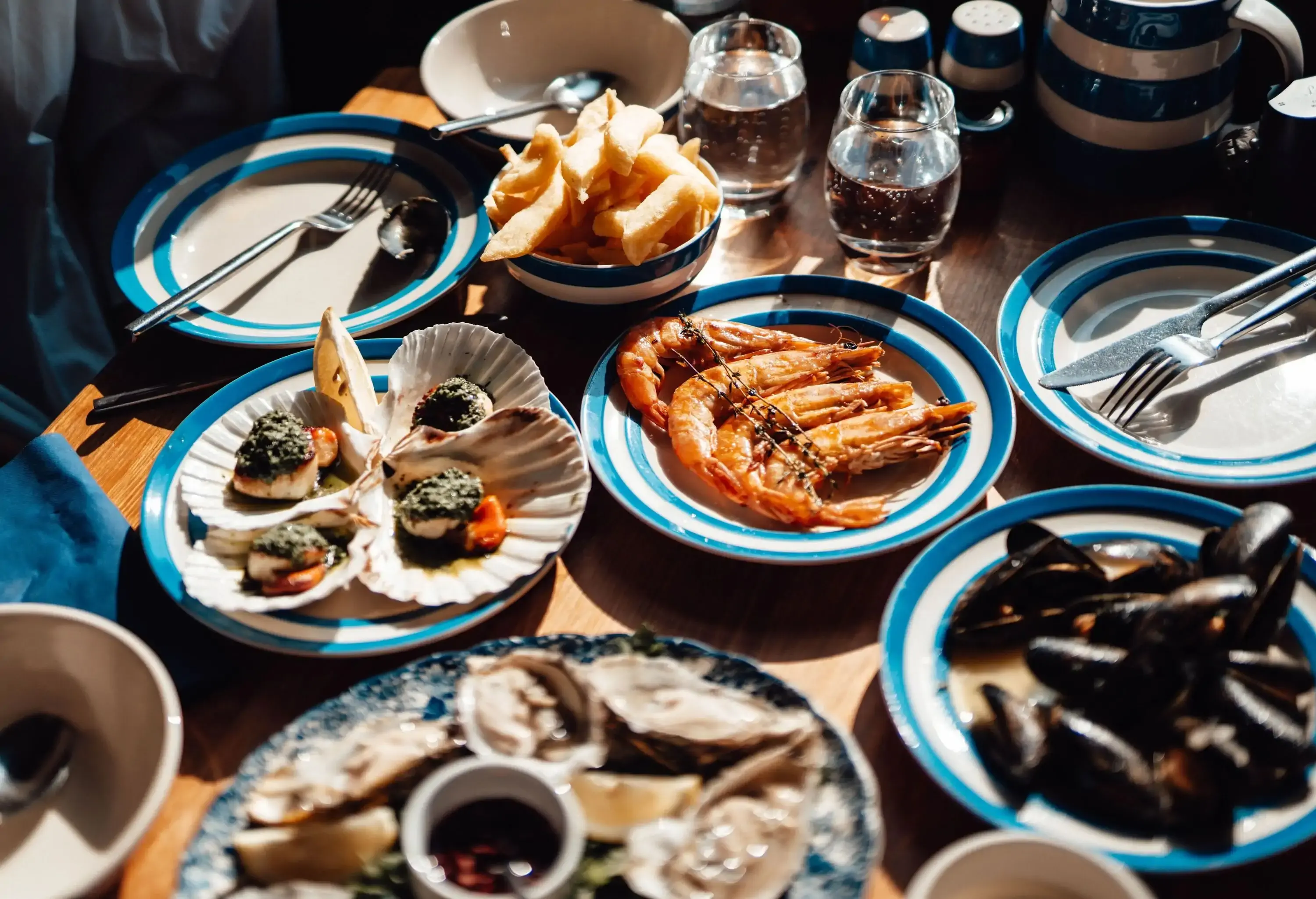 A captivating spread of freshly served seafood, featuring plump oysters, tender scallops, succulent prawns, and briny mussels, is beautifully arranged on a rustic wooden dining table, basking in the warm glow of sunlight.
