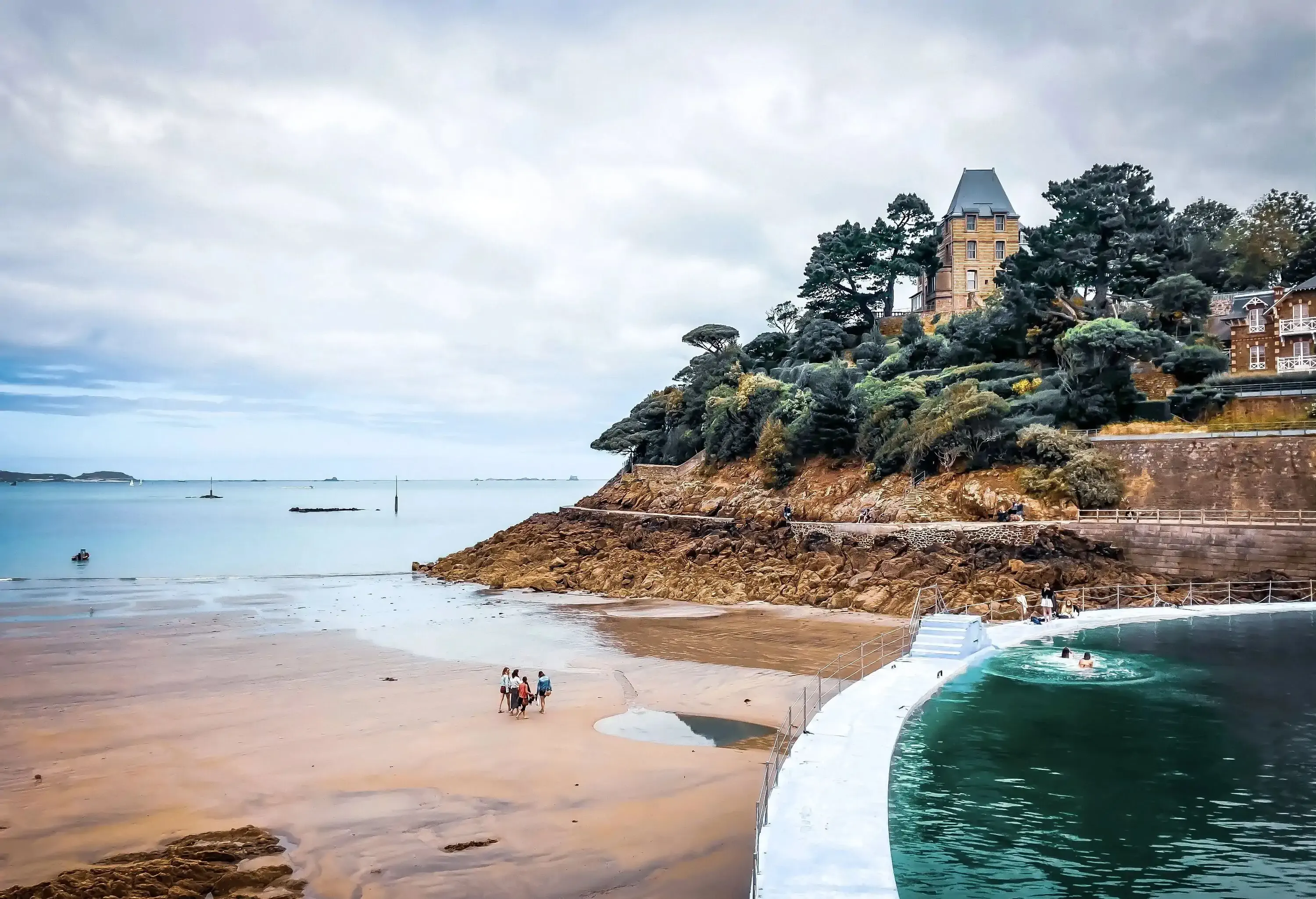 A round pool on the shore of the beach overlooking a classic stone building on top of a nearby lush cliff.