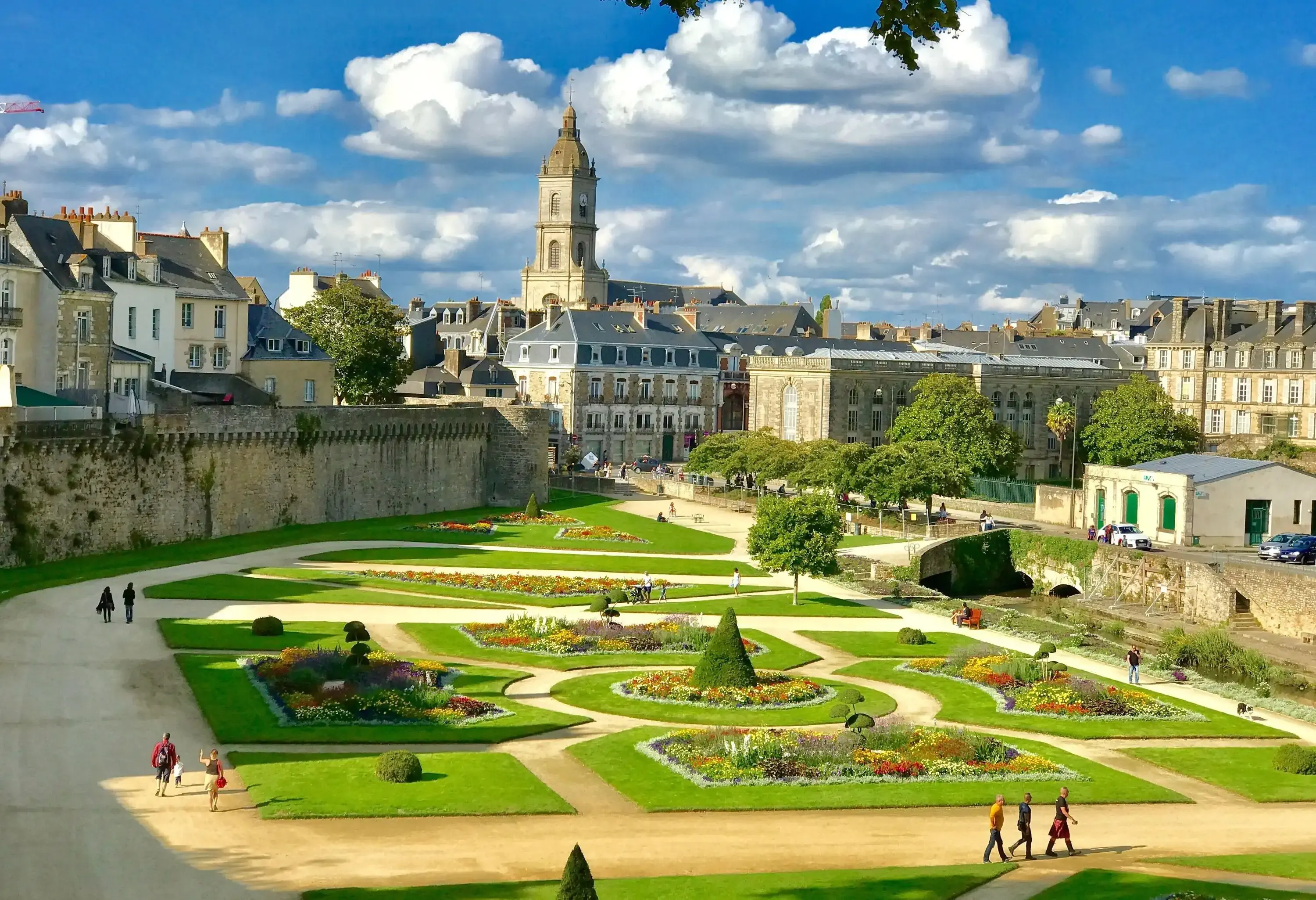 A plaza, where people wander, is bordered by buildings and has grass arranged in different geometric forms with diverse plants in the centre.