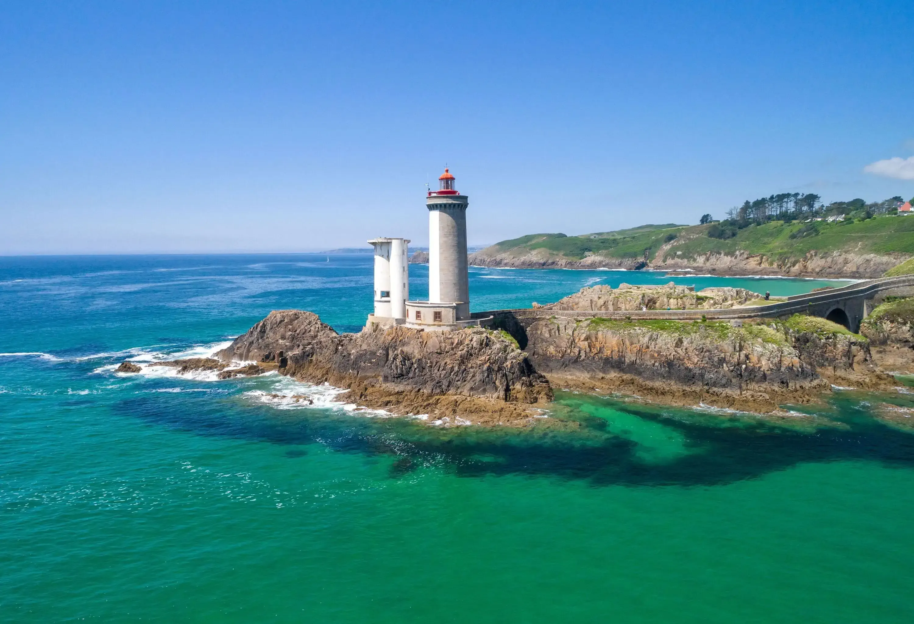 A lighthouse crowned on a rock linked to an arch bridge surrounded by crystal clear waters.