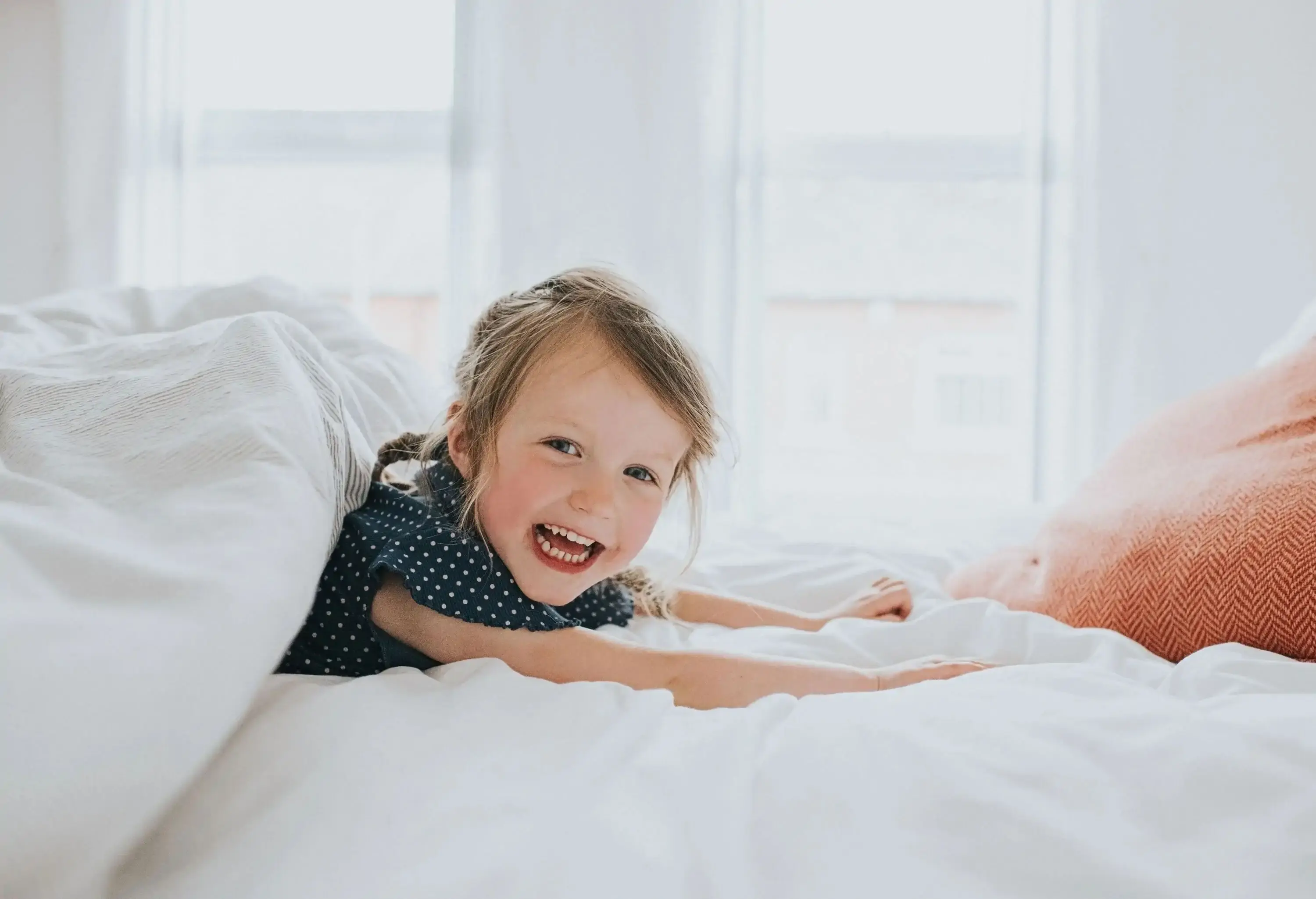 A happy little girl lying on her stomach on a bed with half of her body covered in a white blanket.