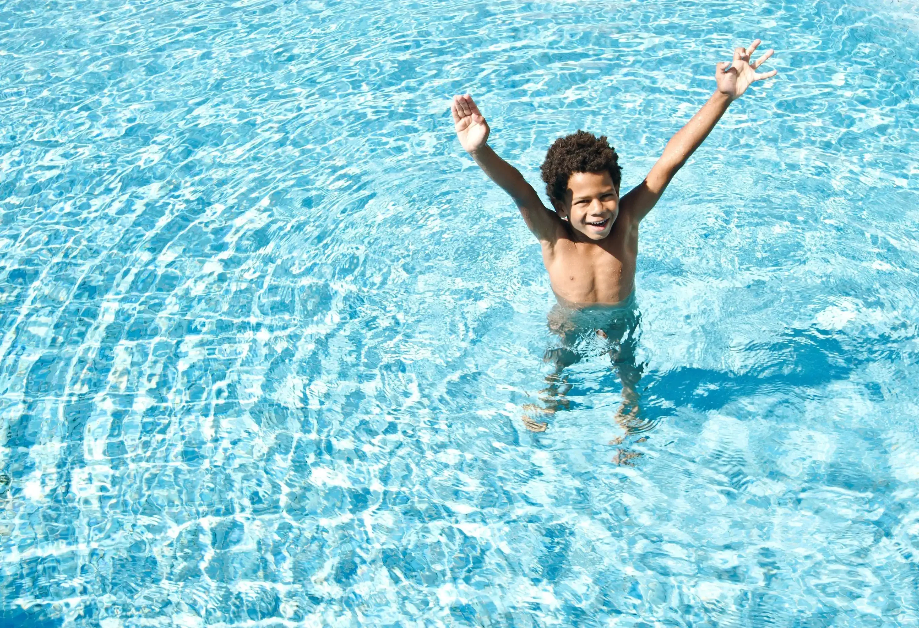 A kid stands with his hands raised on the dazzling swimming pool with water ripples.