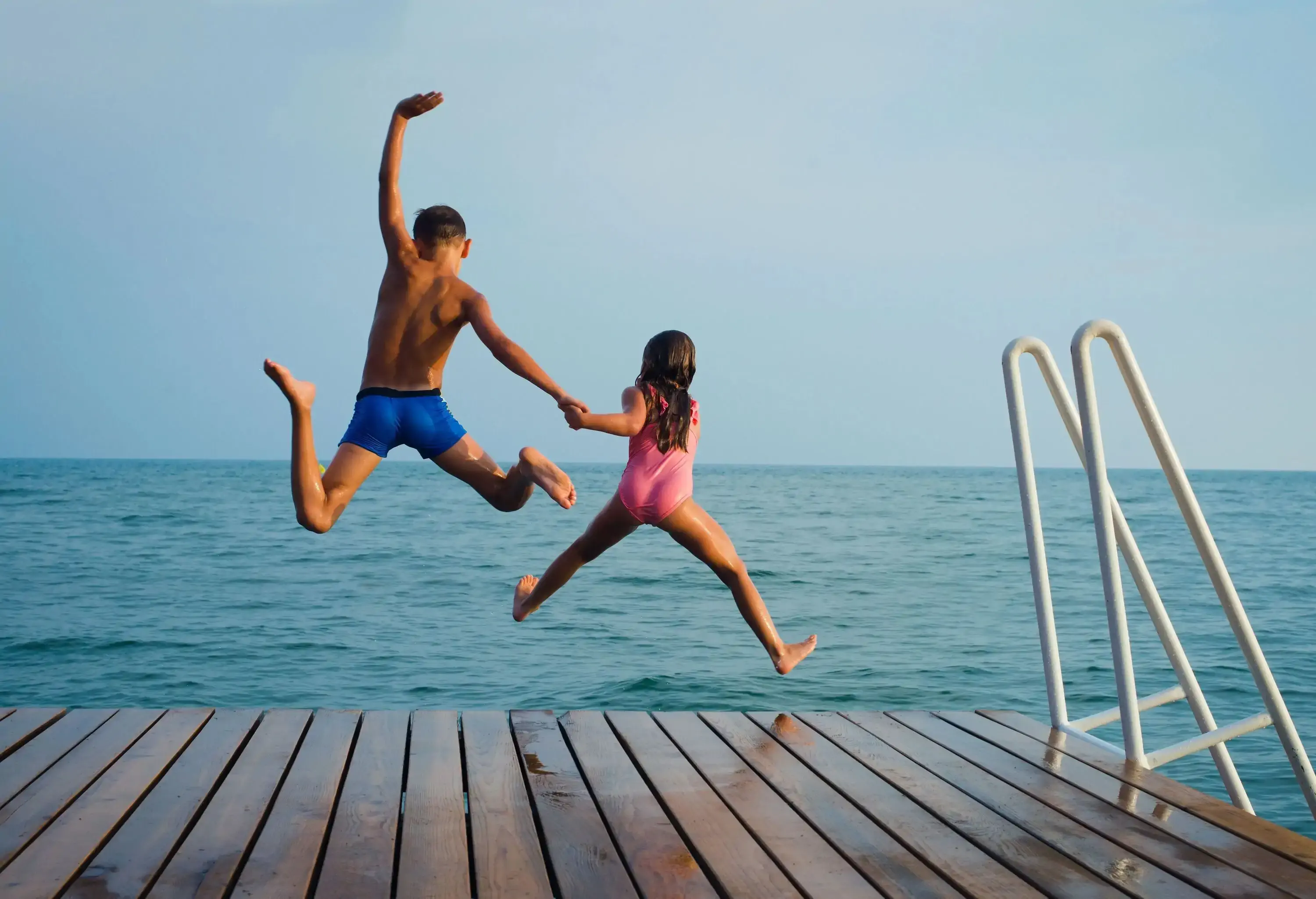 Two children in bathing suits jump into the sea from a wooden platform.