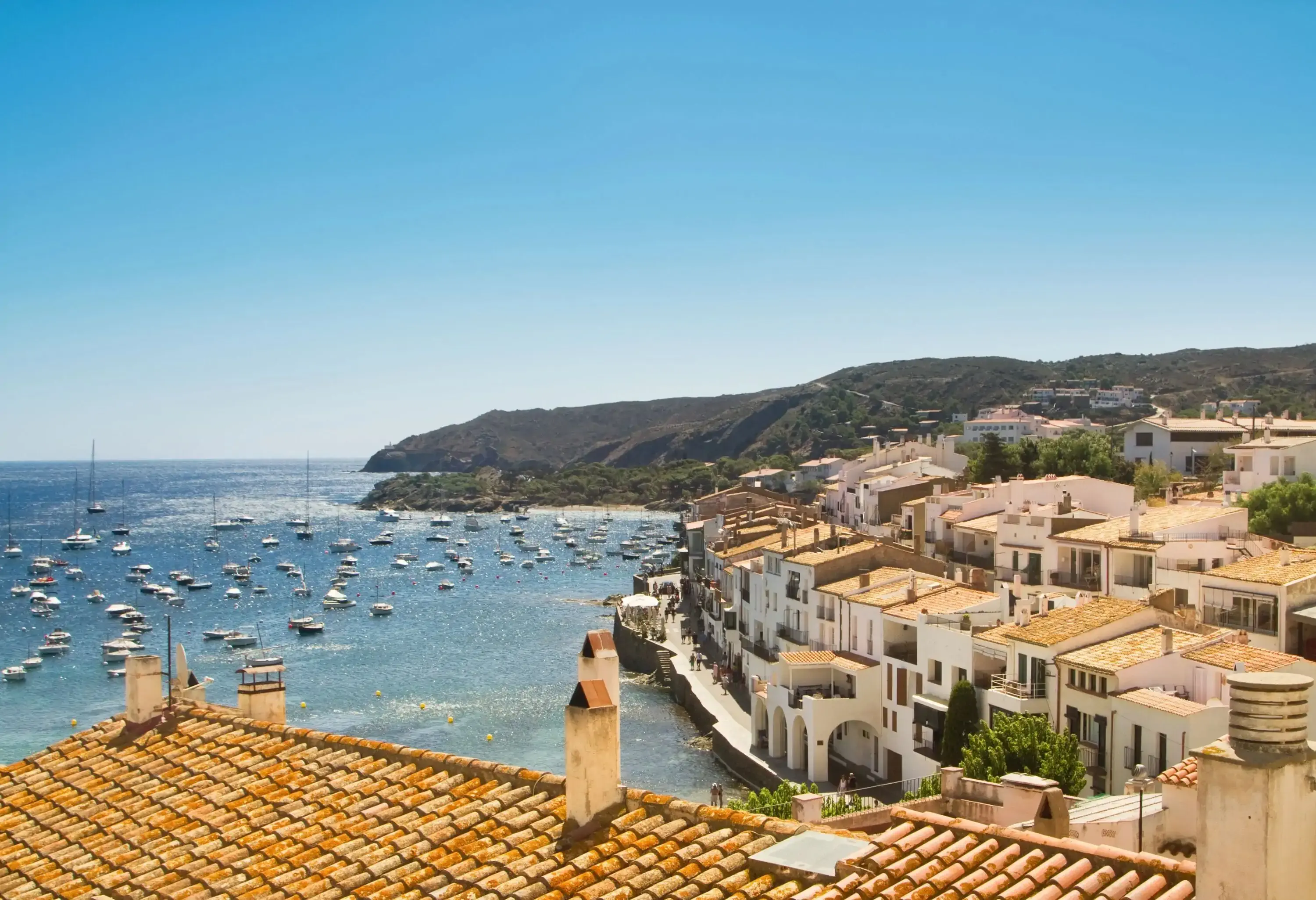 Adjacent town buildings with tiled roofs next to an ocean packed with sailing boats.