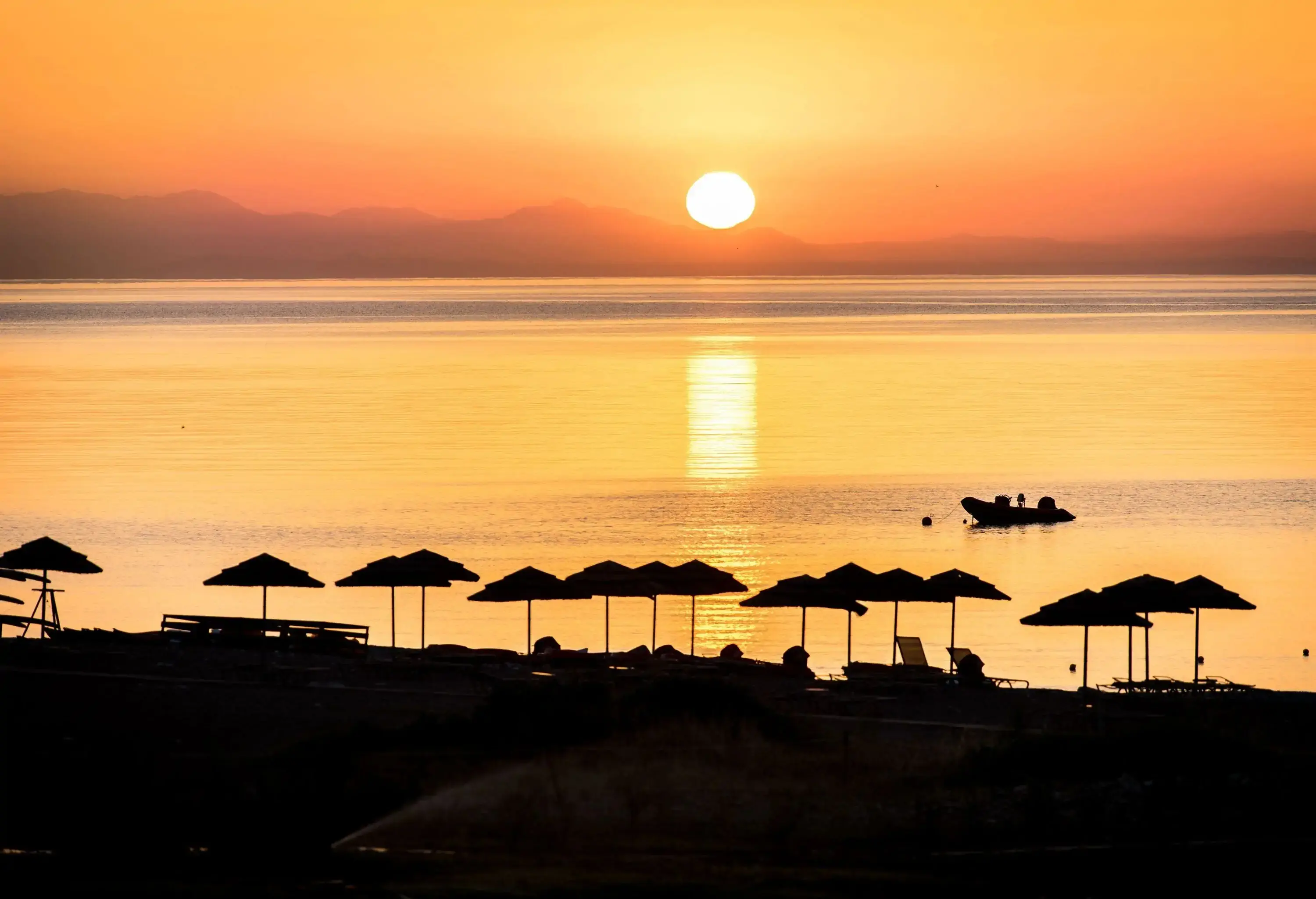 A silhouette of a moored boat and beach umbrellas along the shore of a tranquil sea at sunset.