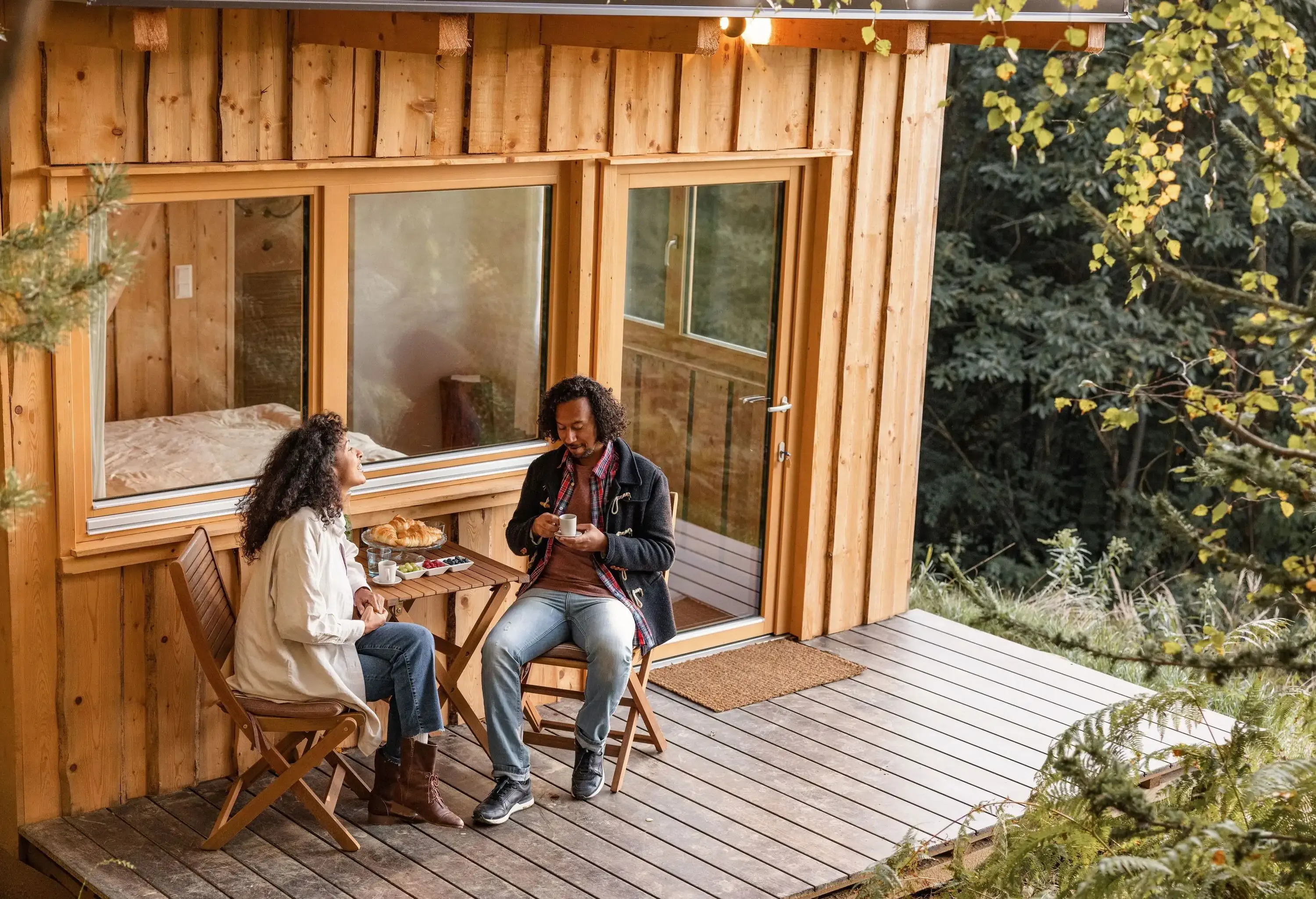 A cosy couple dressed in warm clothes relish their breakfast while sitting on the terrace of a charming wooden cabin.