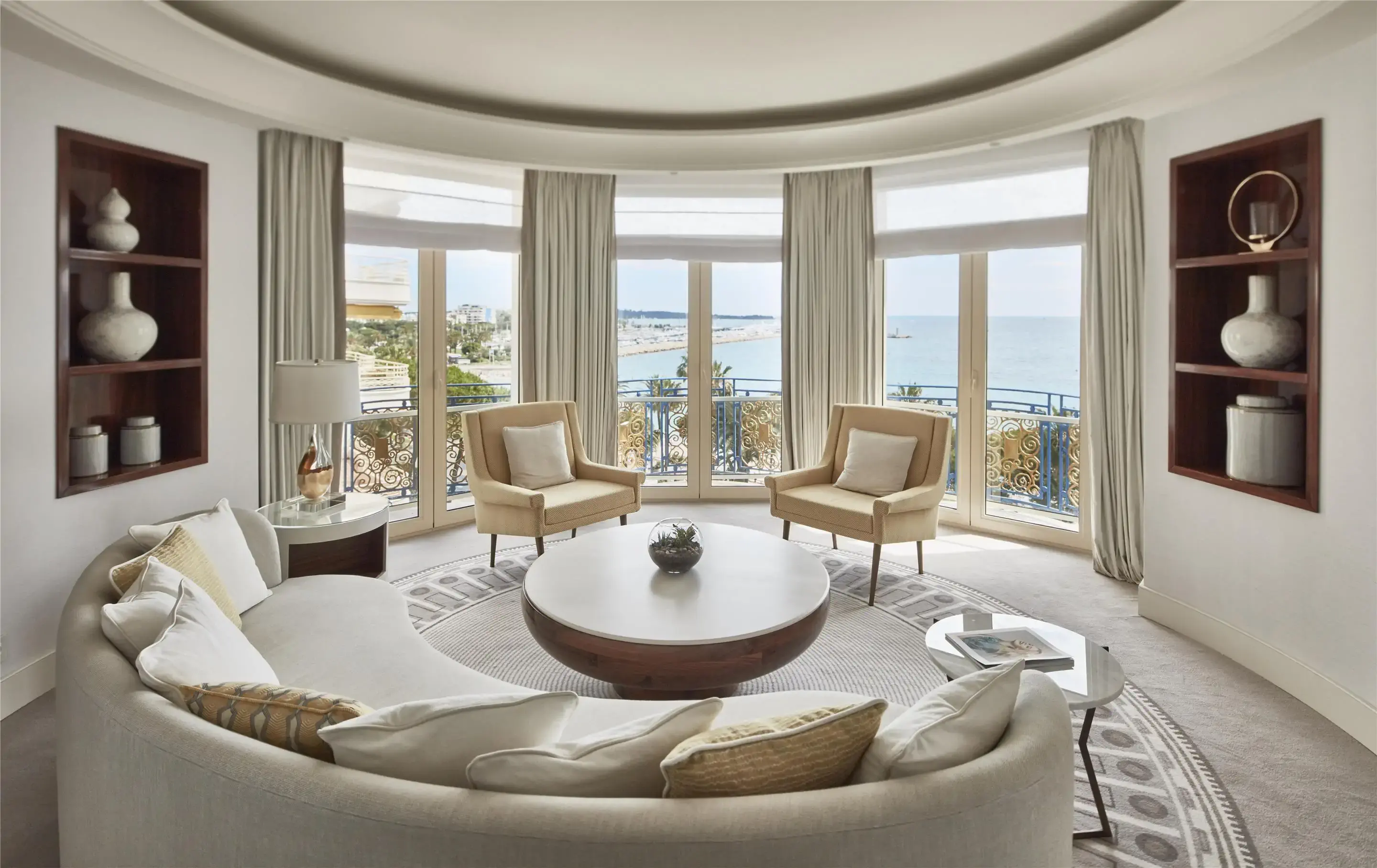 A luxury hotel room built in a circle with view over a bay, two armchairs by the windows and a semi circular sofa in neutral colours
