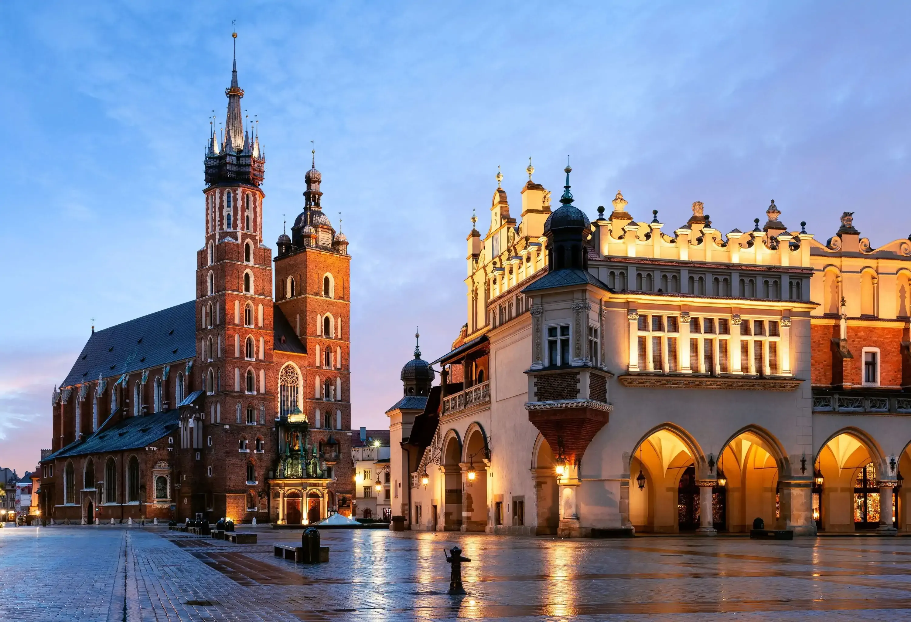 A night-time view of an empty public square along lit buildings with pillared archways and a basilica with towering spires.