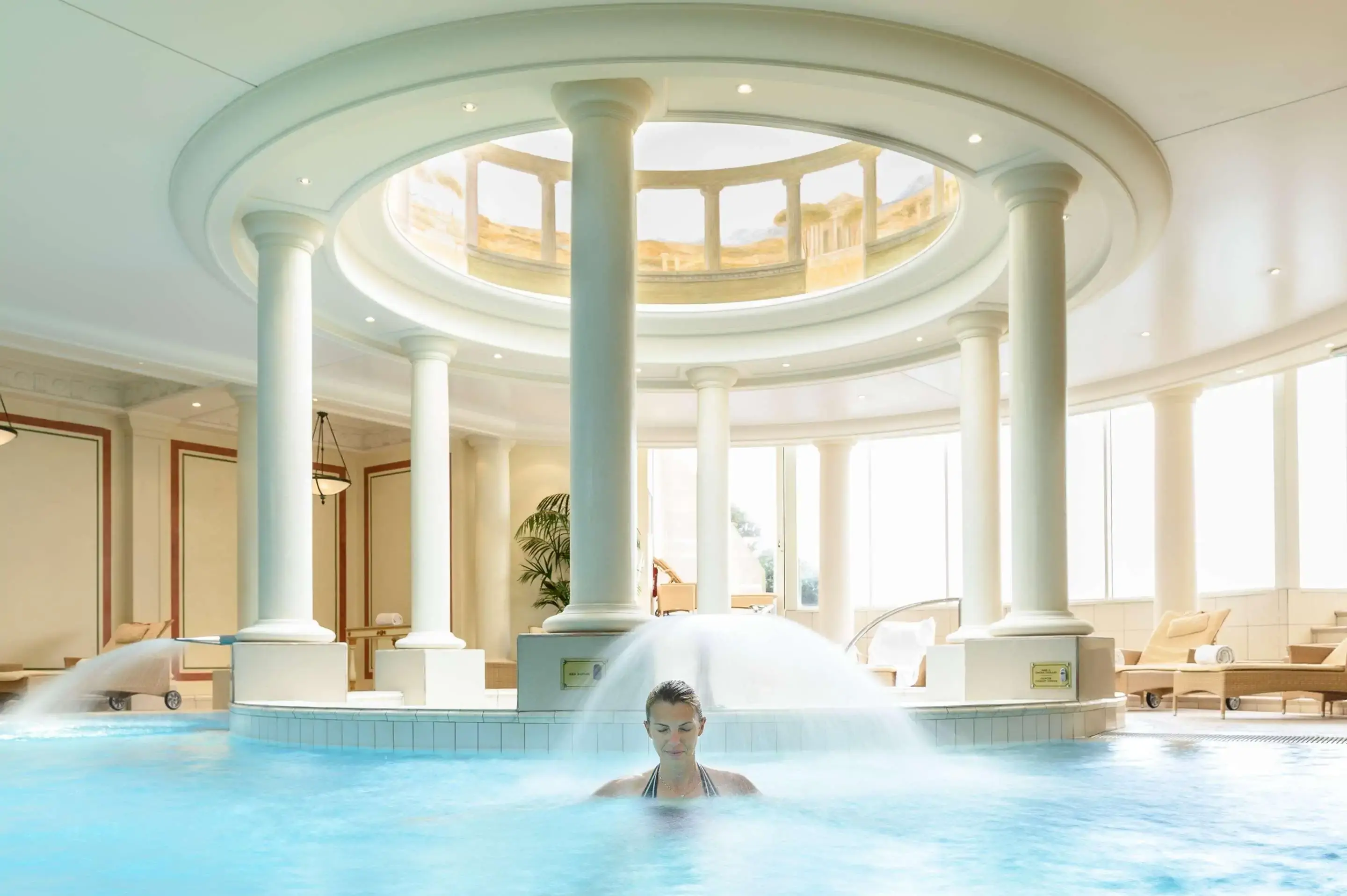 An indoor spa pool with a circular skylight sustained by neoclassical columns and a woman in the water
