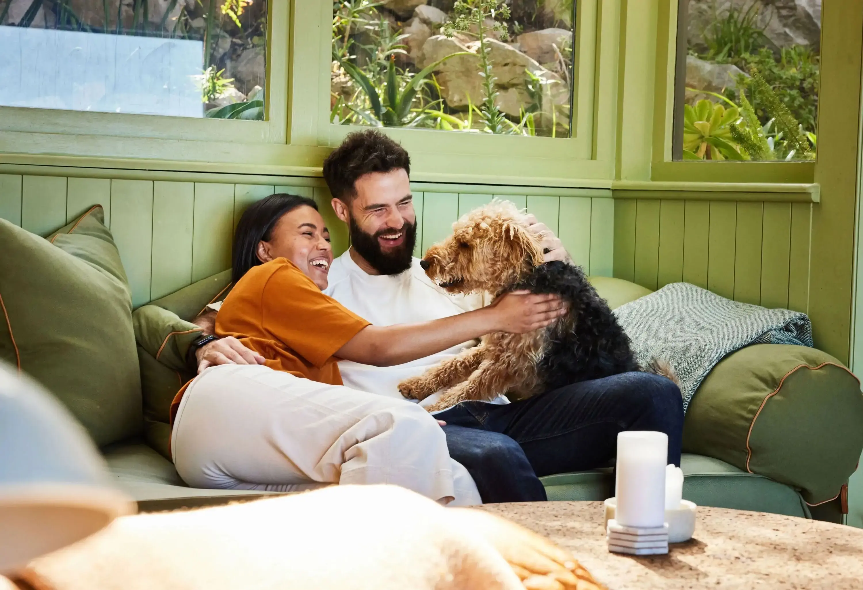 Laughing young couple petting their dog while sitting together on a sofa in their living room at home