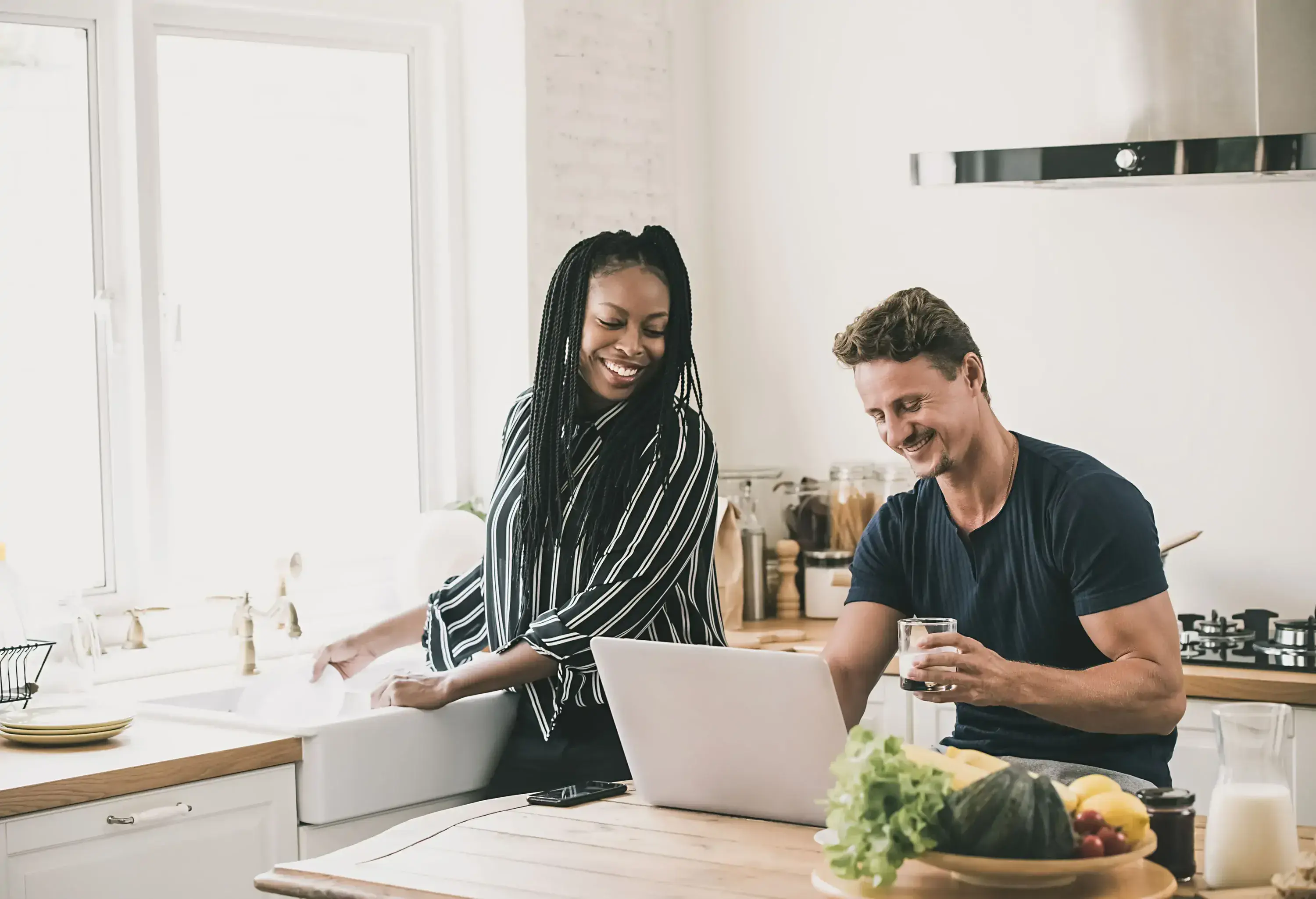 A happy couple in the kitchen, taking a look at the man's laptop while the girl is washing the dishes.