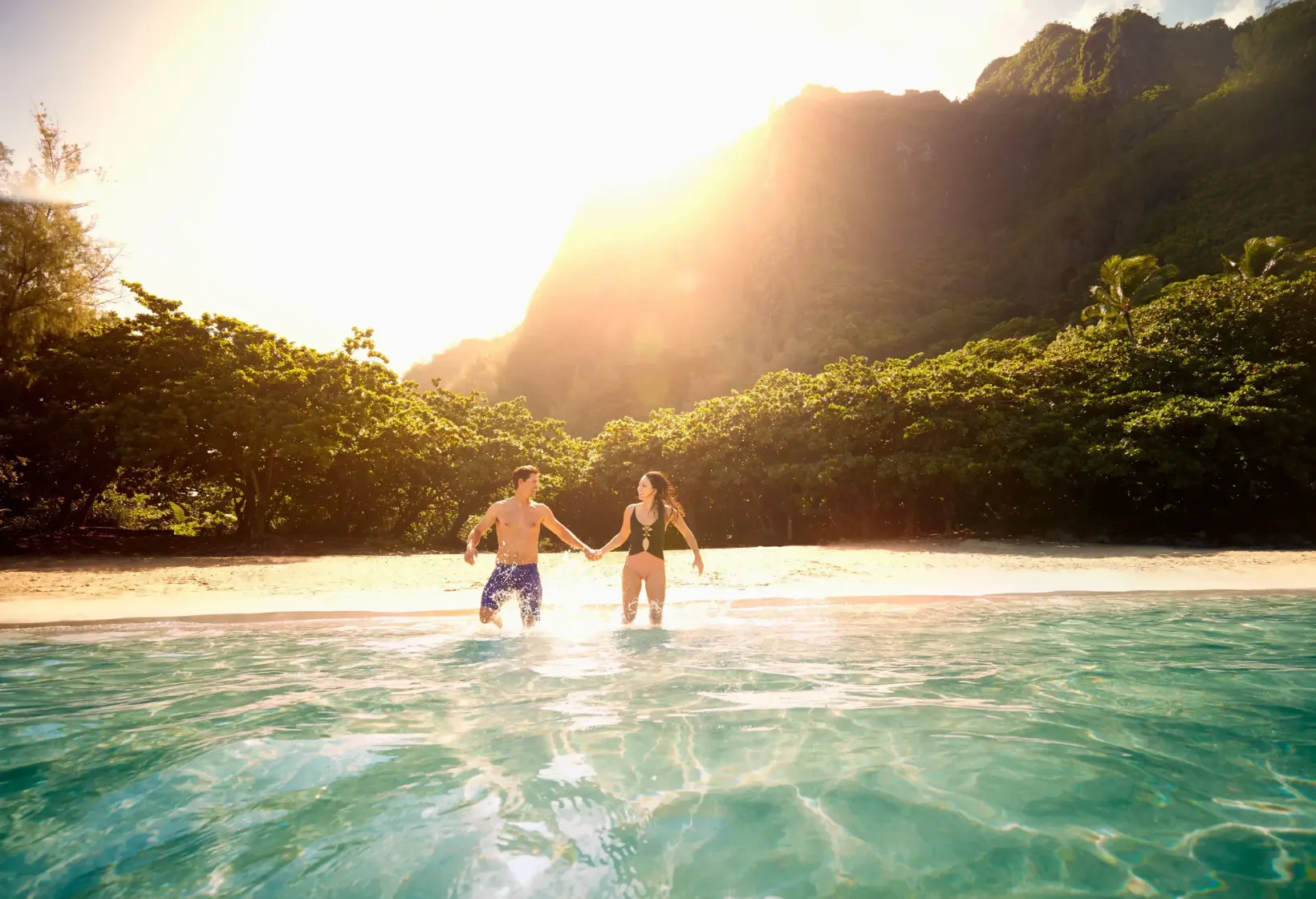 Couple holding hands and walking together into the sparkling clear ocean from a tropical beach, with a bright sun illuminating the distant, forested mountains.