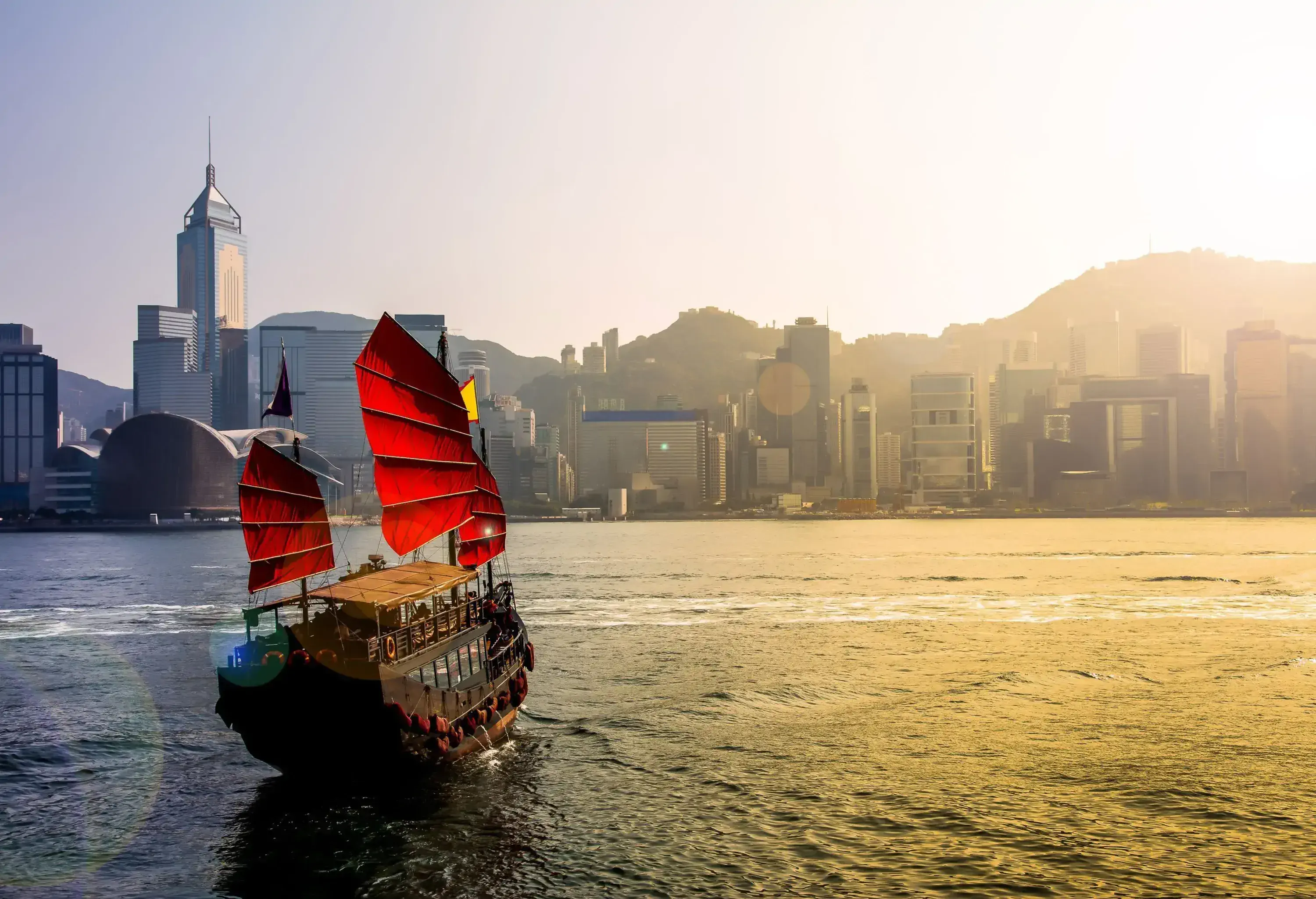 A tourist junk boat sails on the harbour towards the scenic sunlit skyline behind the steep mountains.
