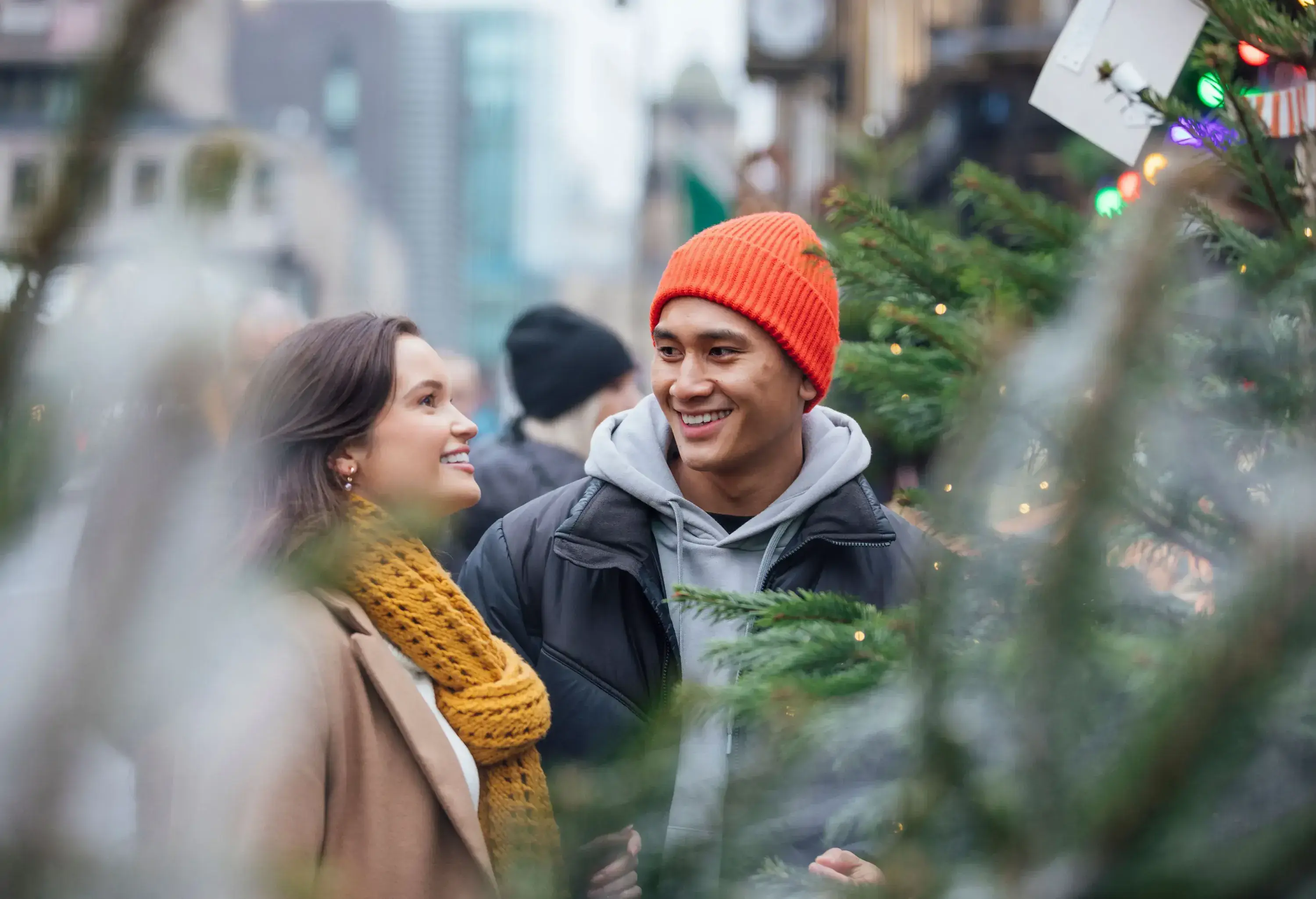 A smiling couple among festive trees adorned with lights in a bustling market.
