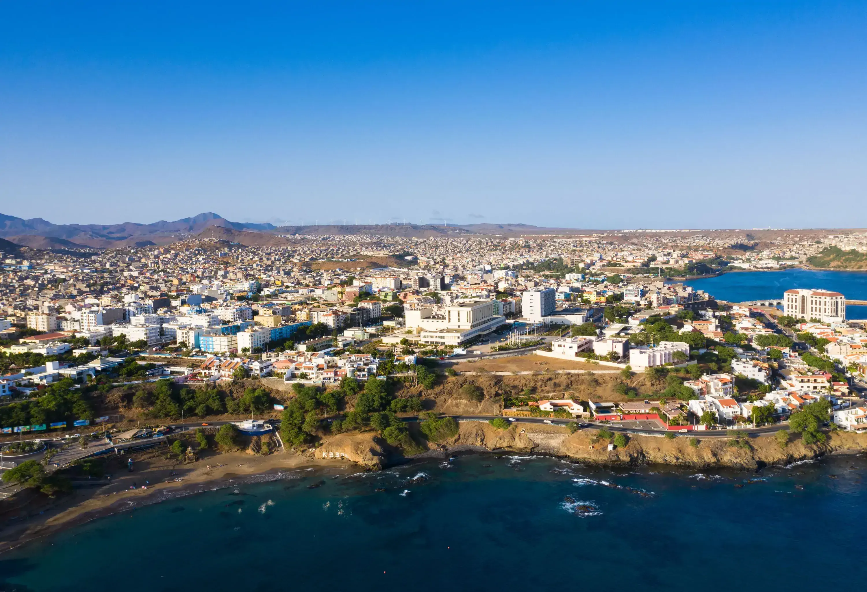 A picturesque city by the coast, where a road lined with trees runs alongside the scenic coastline, featuring buildings that overlook the sea.