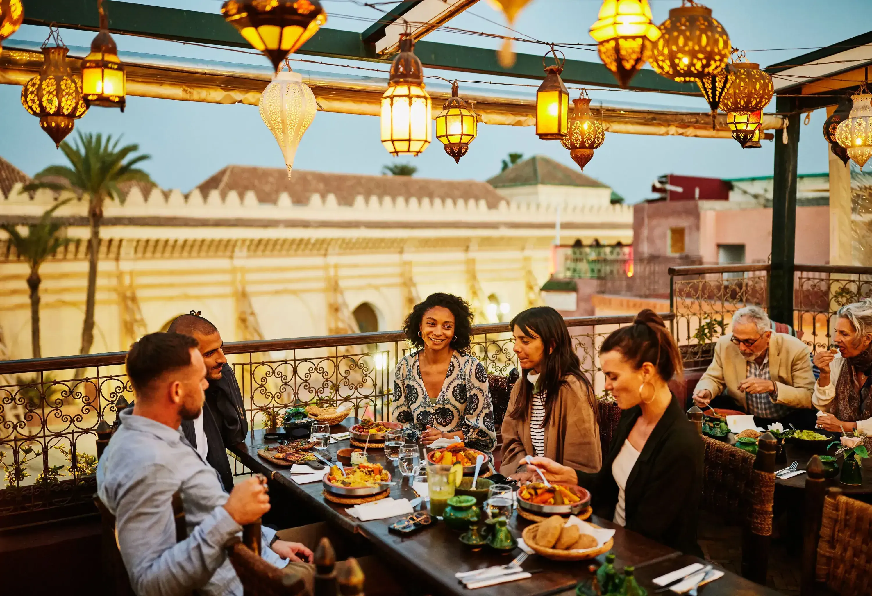 Wide shot of tourists laughing and savoring traditional Moroccan tajine at scenic rooftop eatery during holiday in Marrakech at night