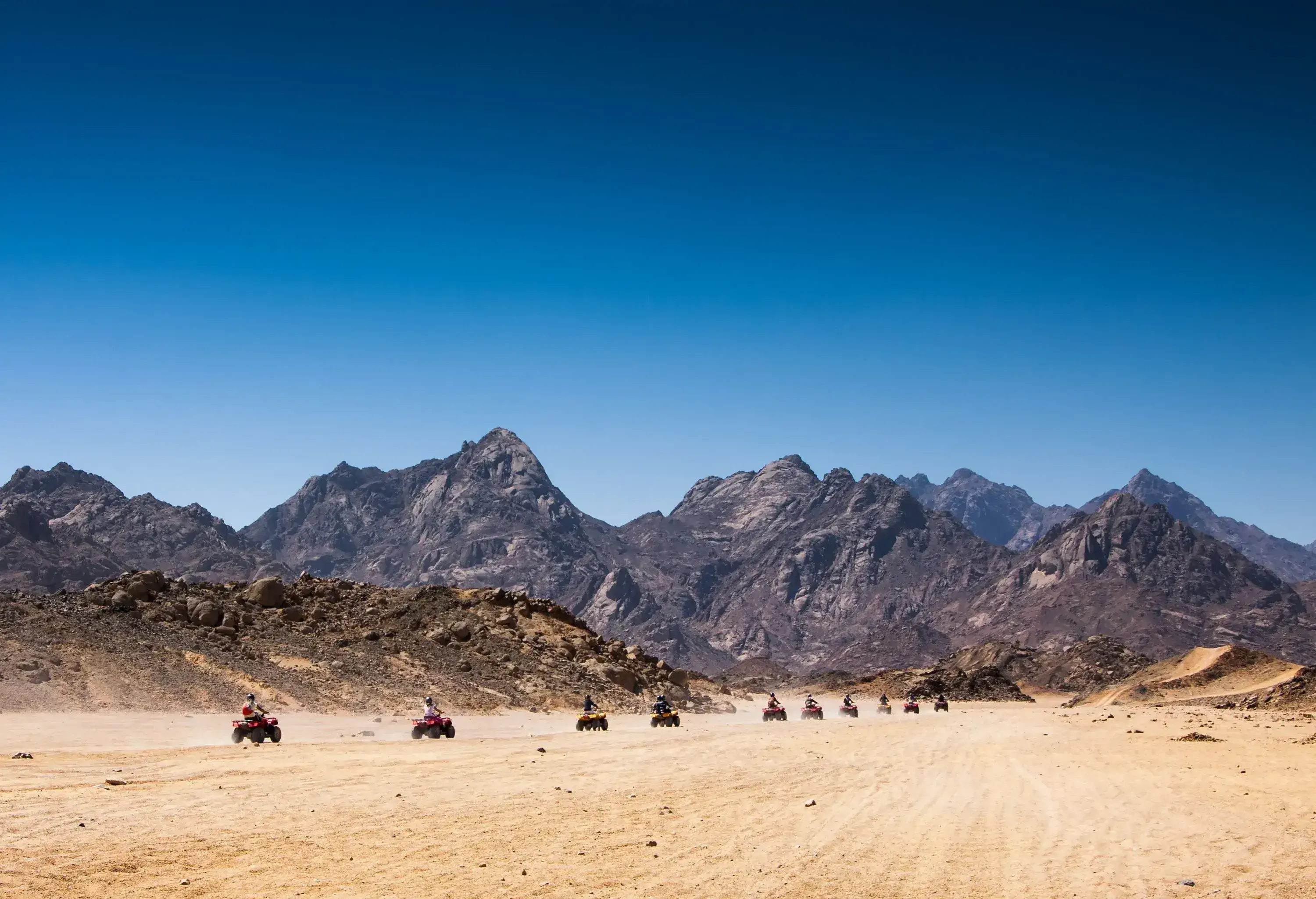 Adventurous tourists riding quad bikes, exploring a vast desert terrain adorned with rocky mountain ranges.