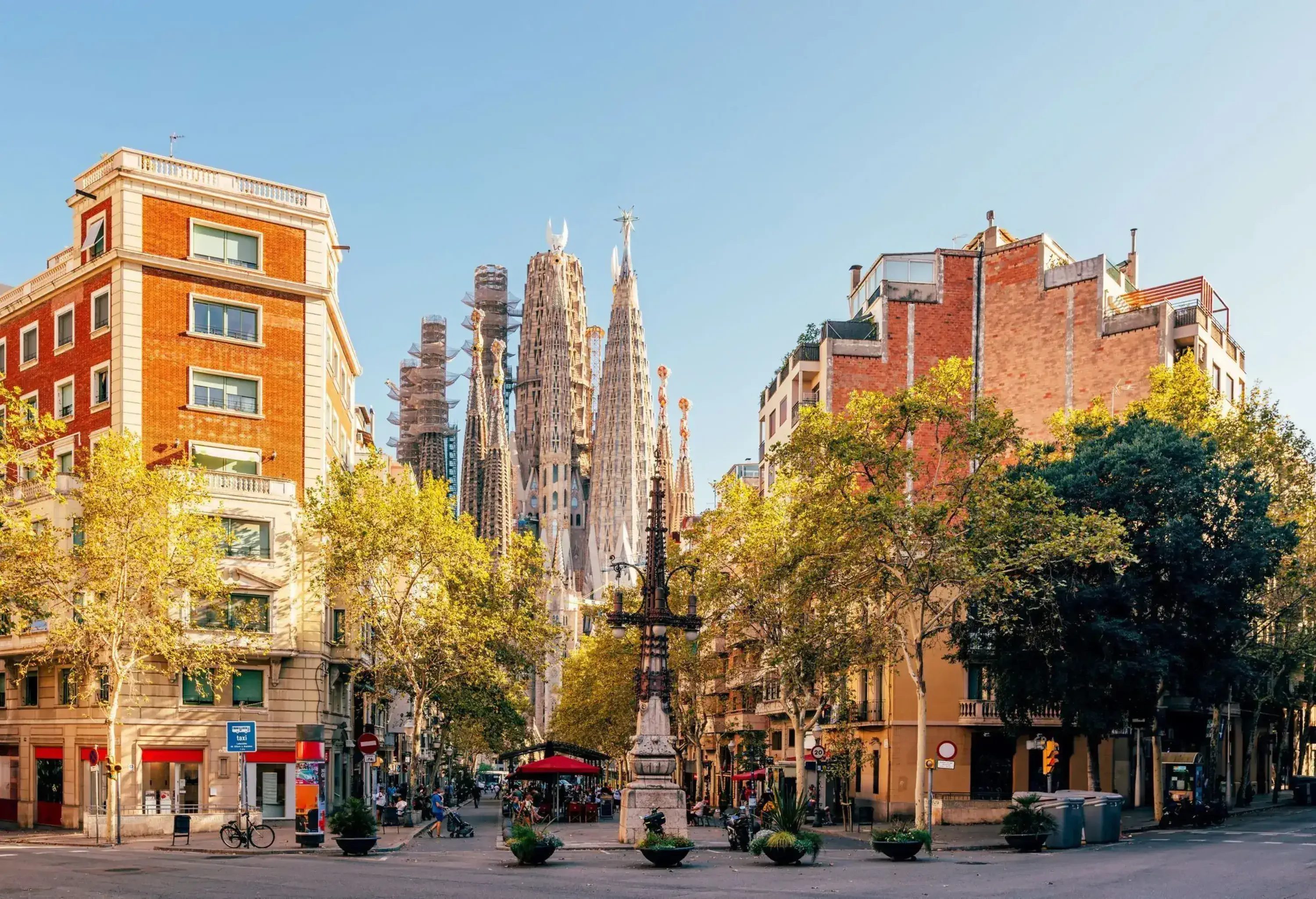 A view of the Sagrada Família basilica under construction, framed by trees, streetlights, and red-brick buildings on a sunny day in Barcelona, Spain.
