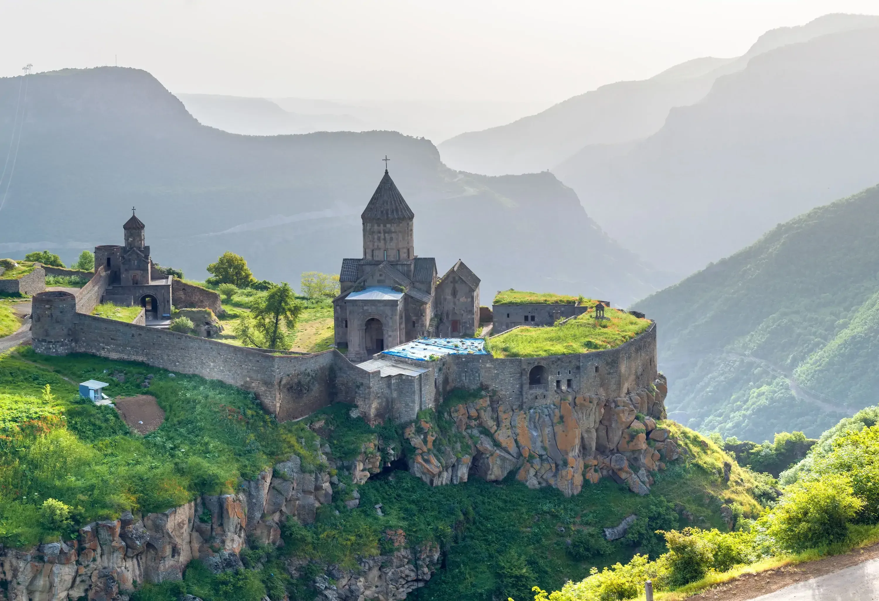 An ancient monastery perches atop a rugged and verdant cliff, its weathered walls hinting at centuries of history, while in the backdrop, a majestic mountainous landscape shrouded in mist adds an aura of mystery and enchantment to the scene.
