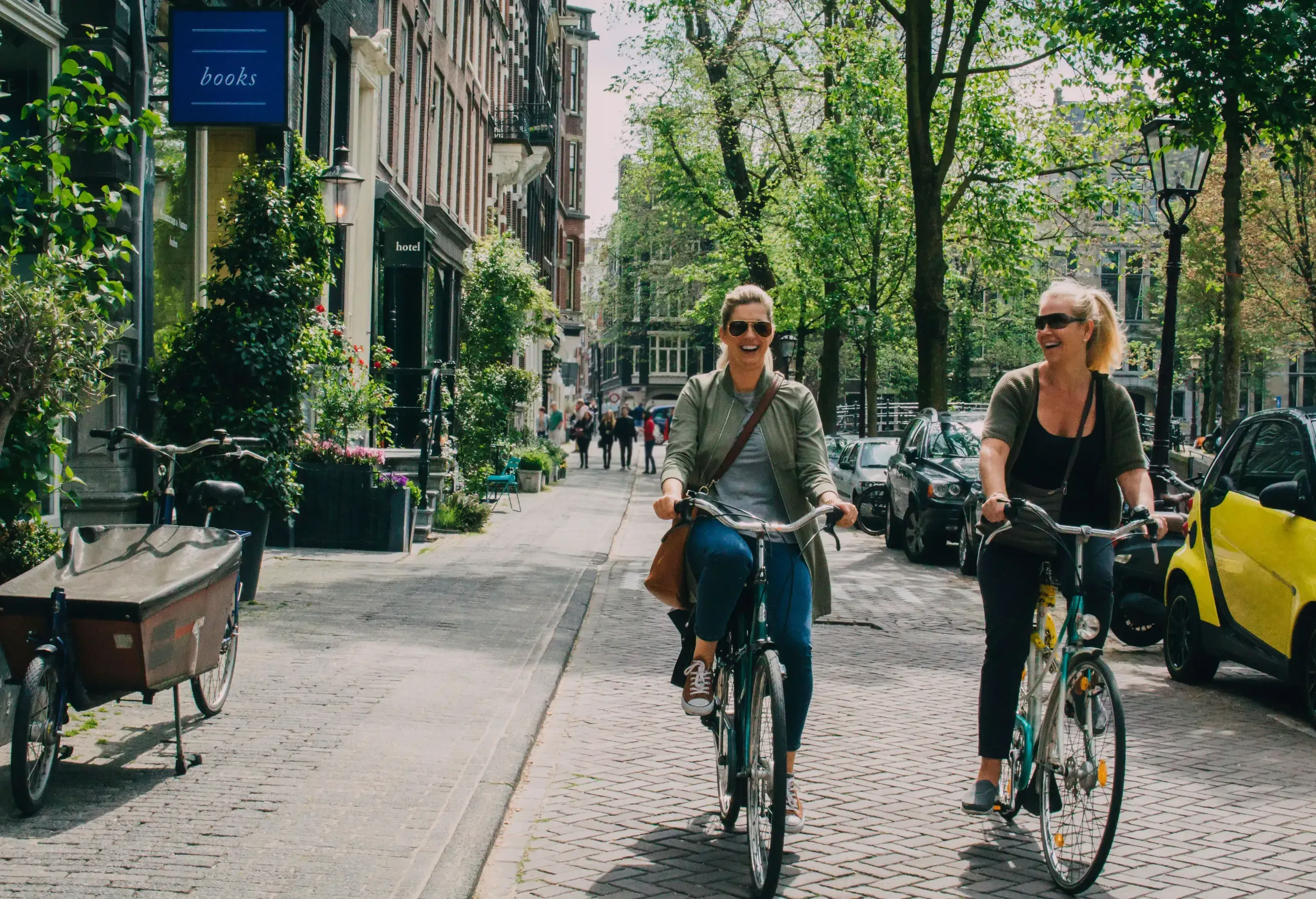 Two women riding their bikes on a pavement lined with trees and cars.